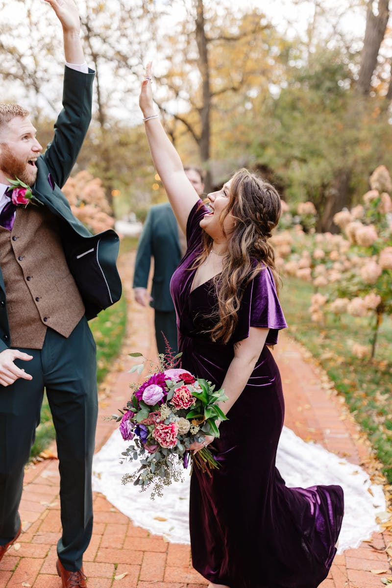 Jewel tone wedding party with groomsman in a dark green suit and brown tweed vest high-fiving bridesmaid in eggplant purple velvet dress.