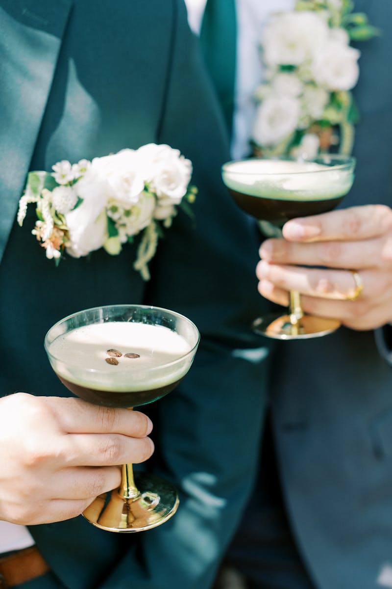 Gay grooms wearing green suit and dark grey suit with big boutonnieres holding espresso martini wedding signature cocktail