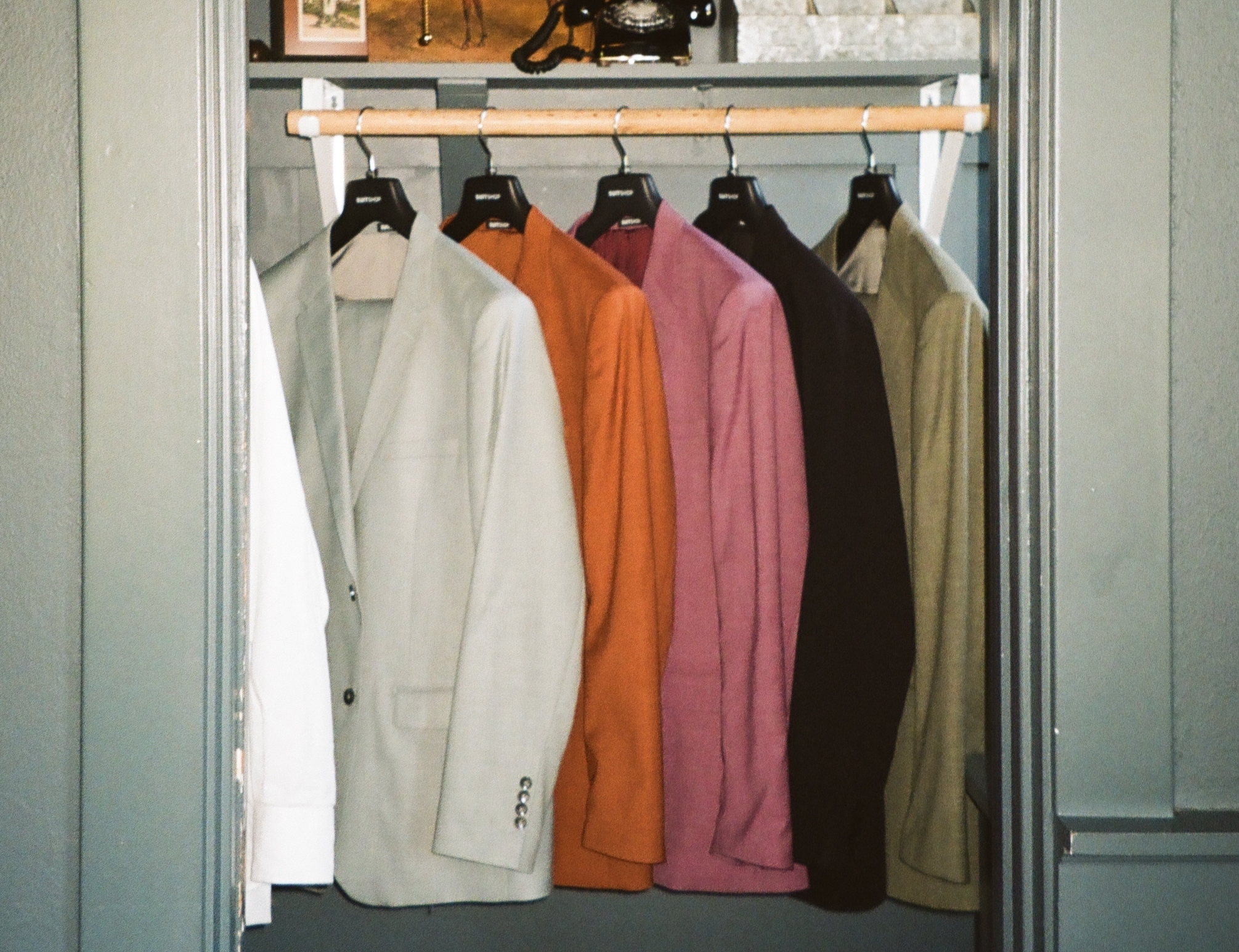 Five mismatched groomsmen suit jackets in cream, burnt orange, dusty pink, dark brown, and olive green hanging side by side in a closet.