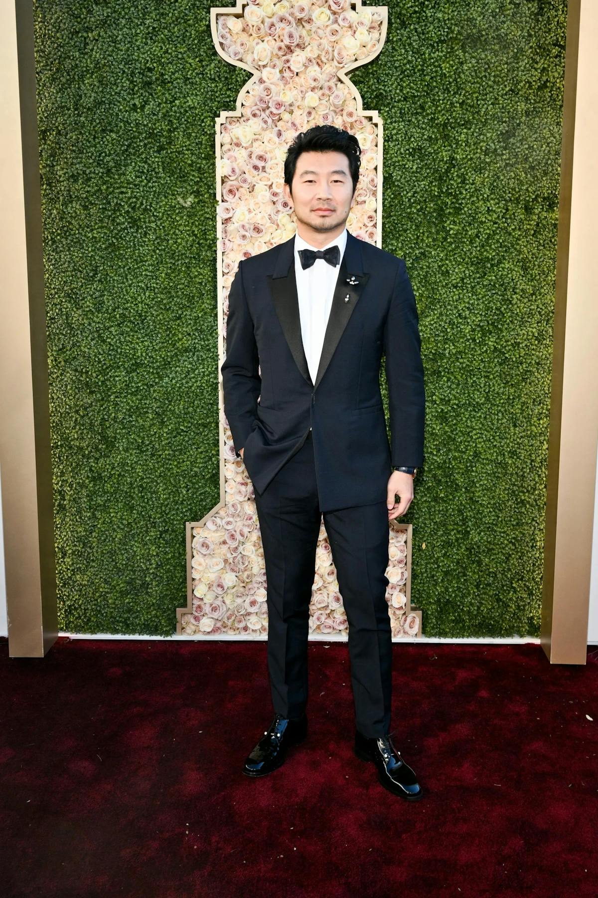 Simu Liu in a peak lapel navy tuxedo jacket with black tuxedo pants and lapel pins at the Golden Globes 2024 red carpet.