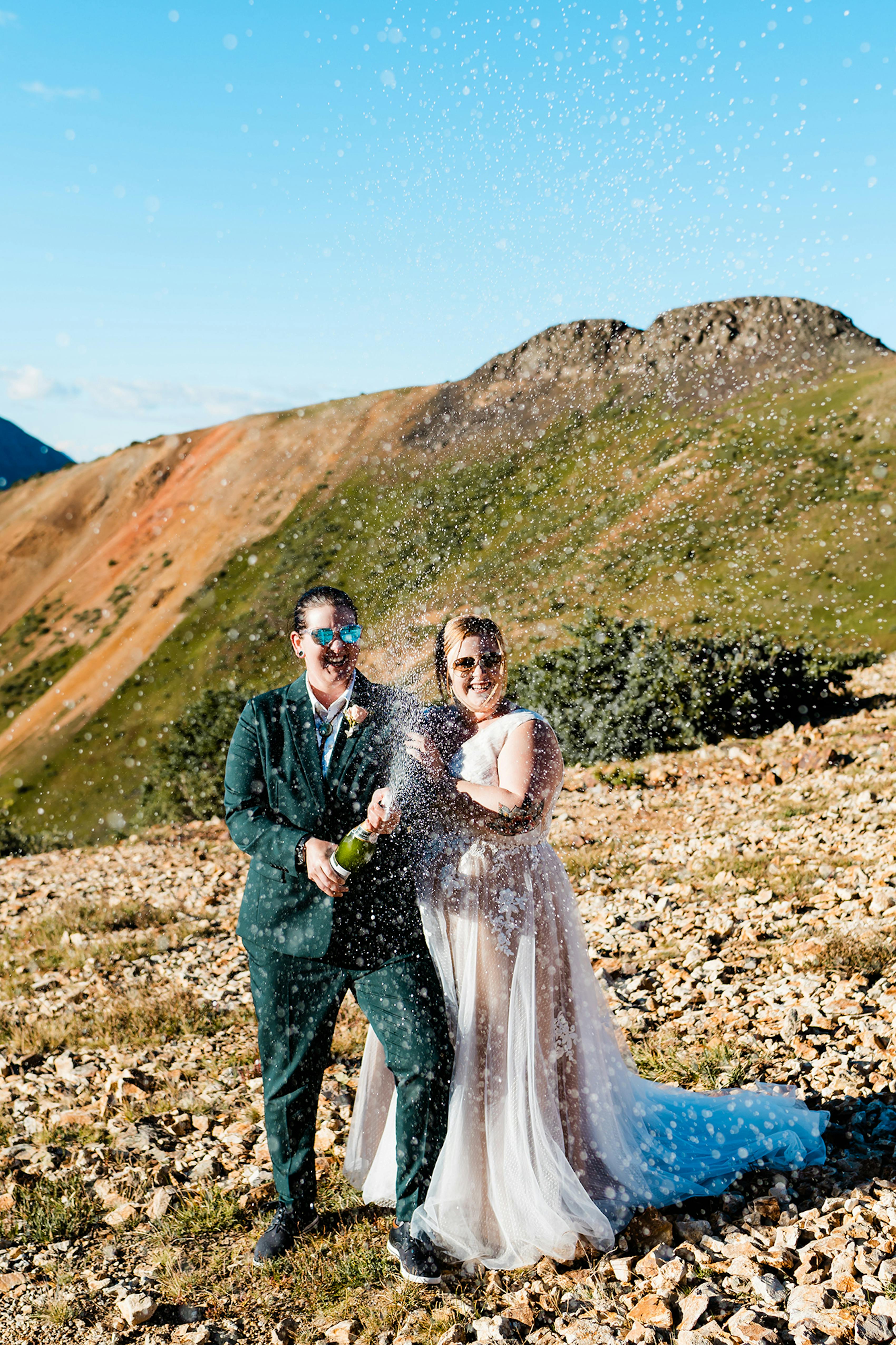 Brides in dark green women's wedding suit and gown popping champagne.