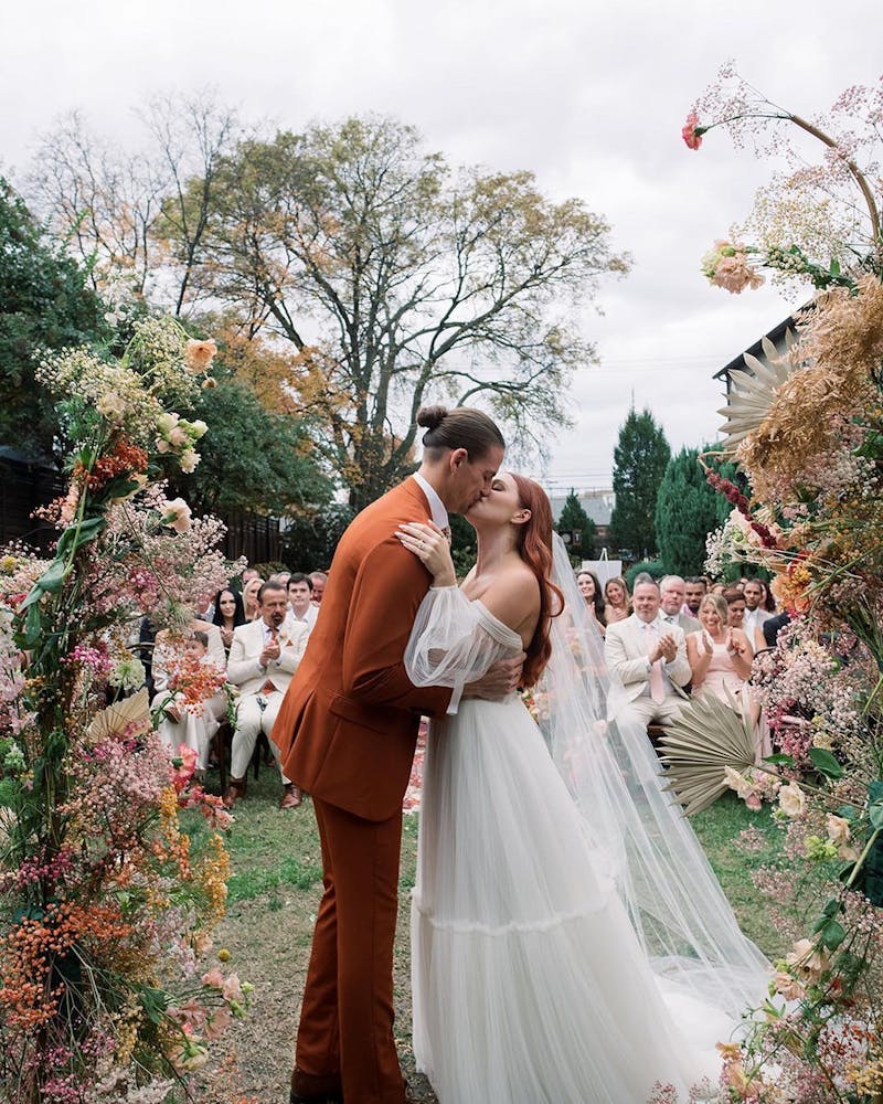 Groom wearing rust men's orange suit and bride wearing boho tulle dress at flower altar in front of neutral themed garden wedding guests