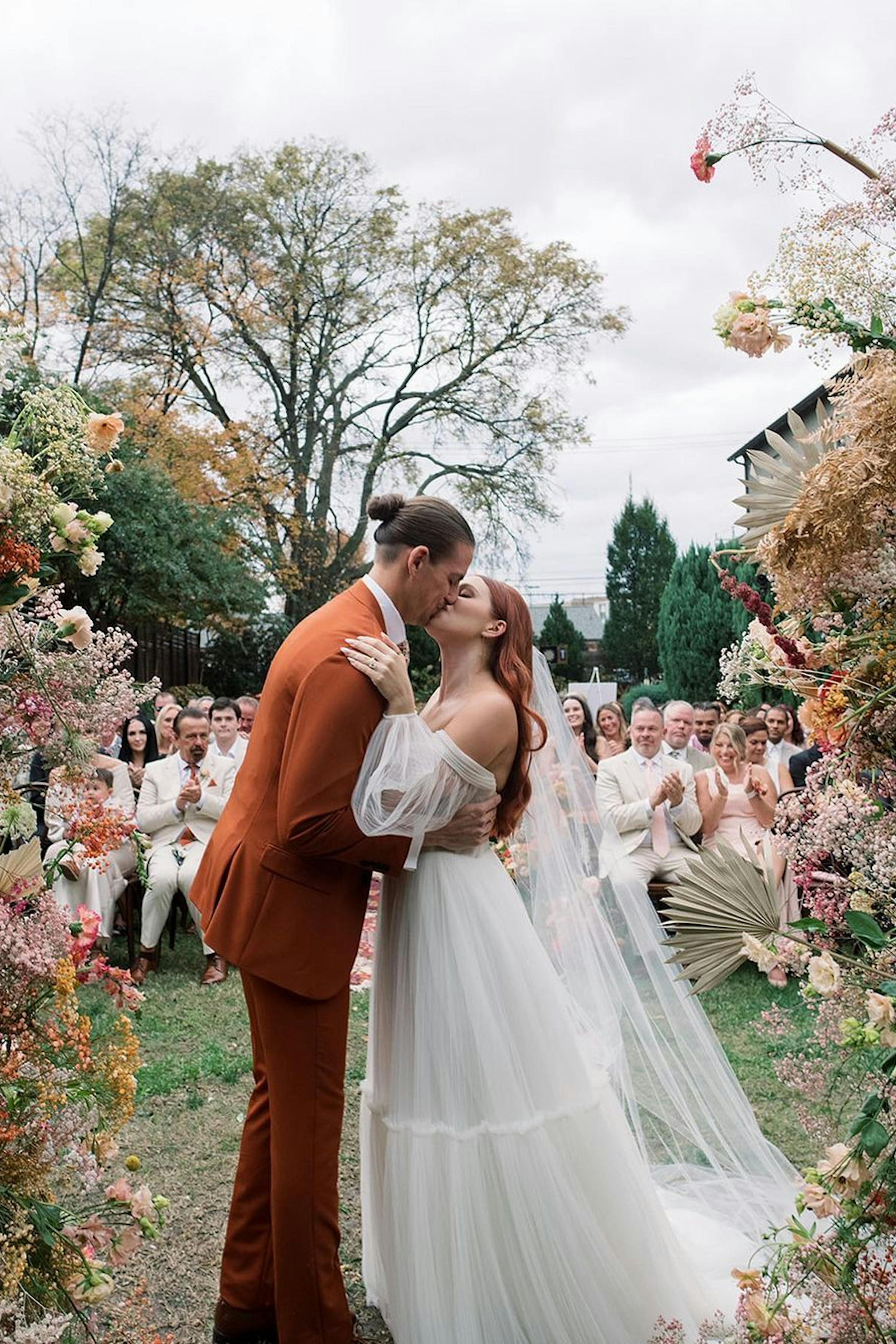 Groom wearing rust men's orange suit and bride wearing boho tulle dress at flower altar in front of neutral themed garden wedding guests