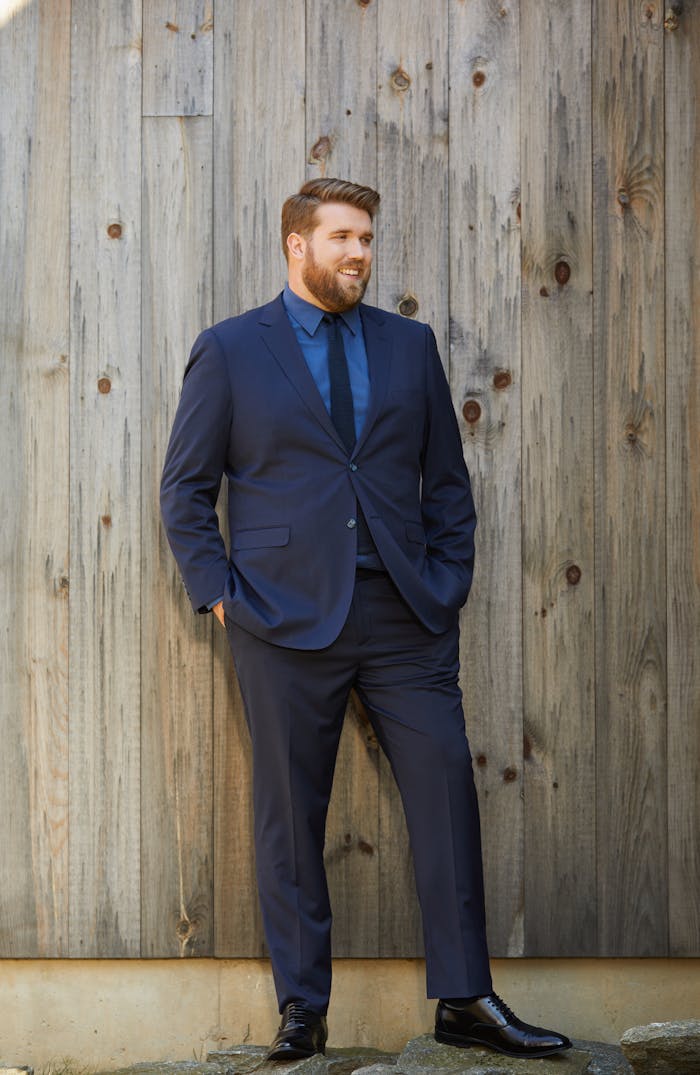 A man in a dark blue suit with a bright blue dress shirt and black suit accessories for a colorful wedding guest outfit to wear for cocktail events.