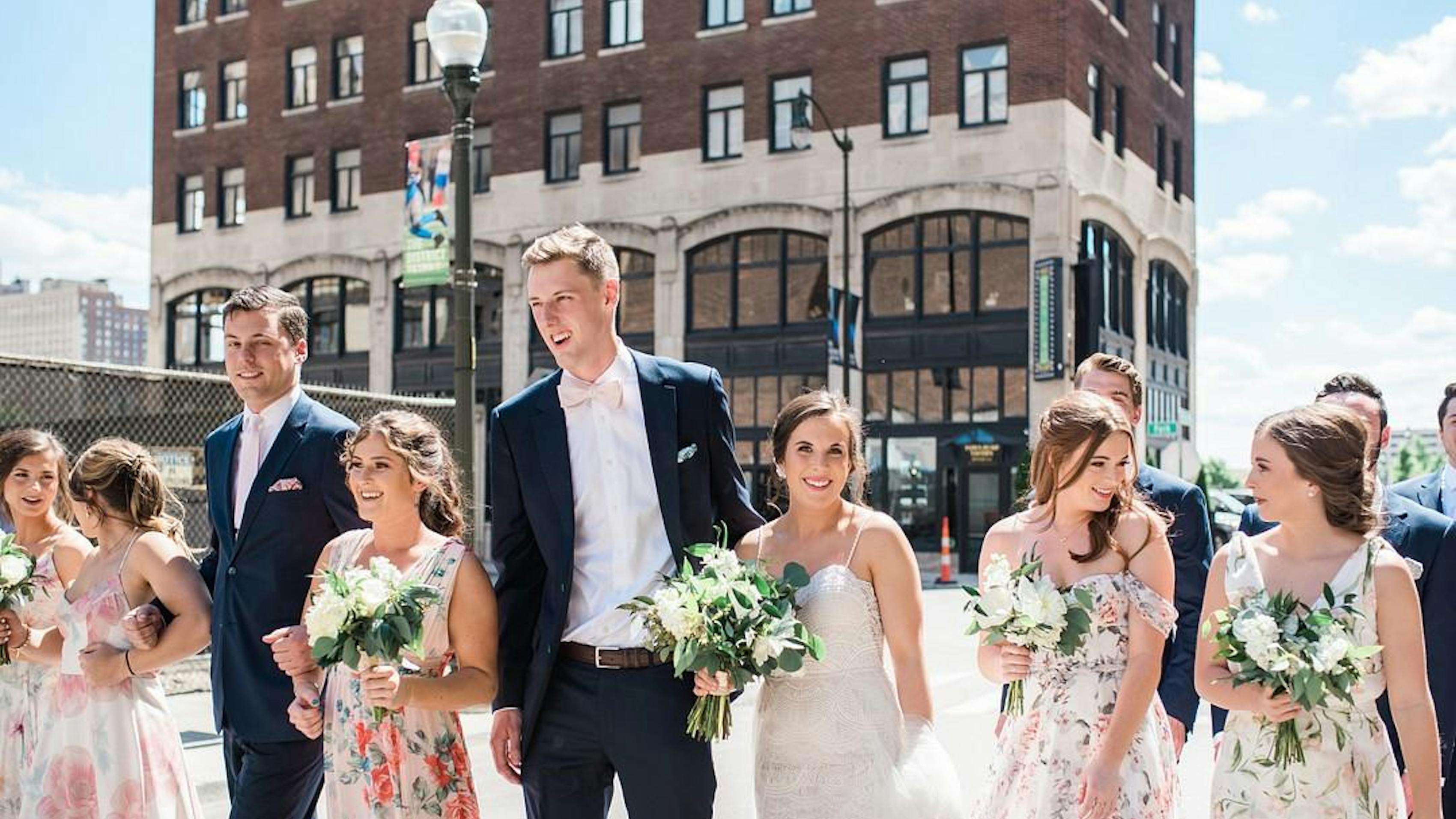 Trendy wedding party in mismatched floral bridesmaid gowns and navy groomsman suits and pink accessories with city background.