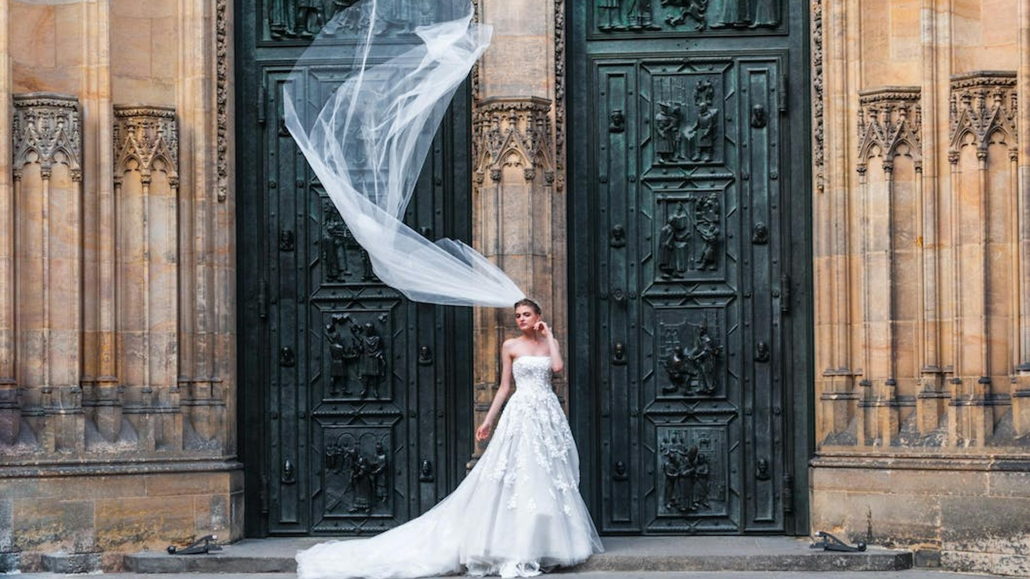 Strapless floral lace wedding gown with full tulle skirt and long veil blowing in the wind in front of traditional church.