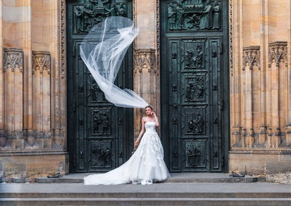 Strapless floral lace wedding gown with full tulle skirt and long veil blowing in the wind in front of traditional church.