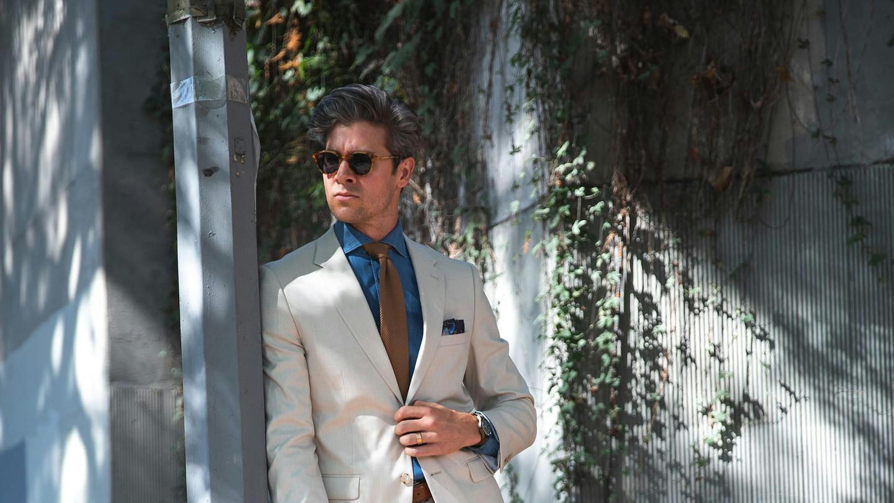 Man outside showing what to wear with a tan suit, including a blue button up shirt, a brown tie, brown socks, and white loafers.