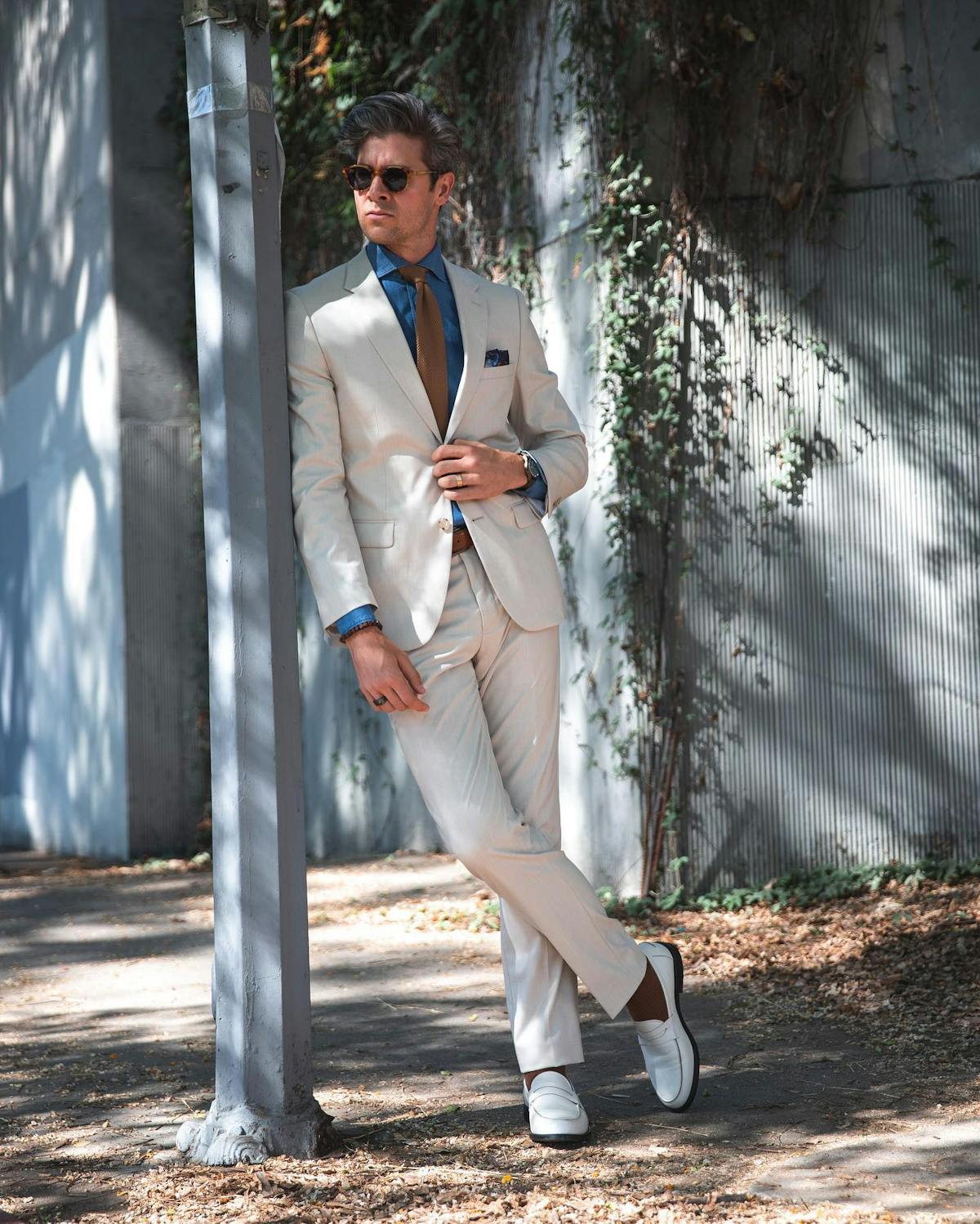 Man outside showing what to wear with a tan suit, including a blue button up shirt, a brown tie, brown socks, and white loafers.