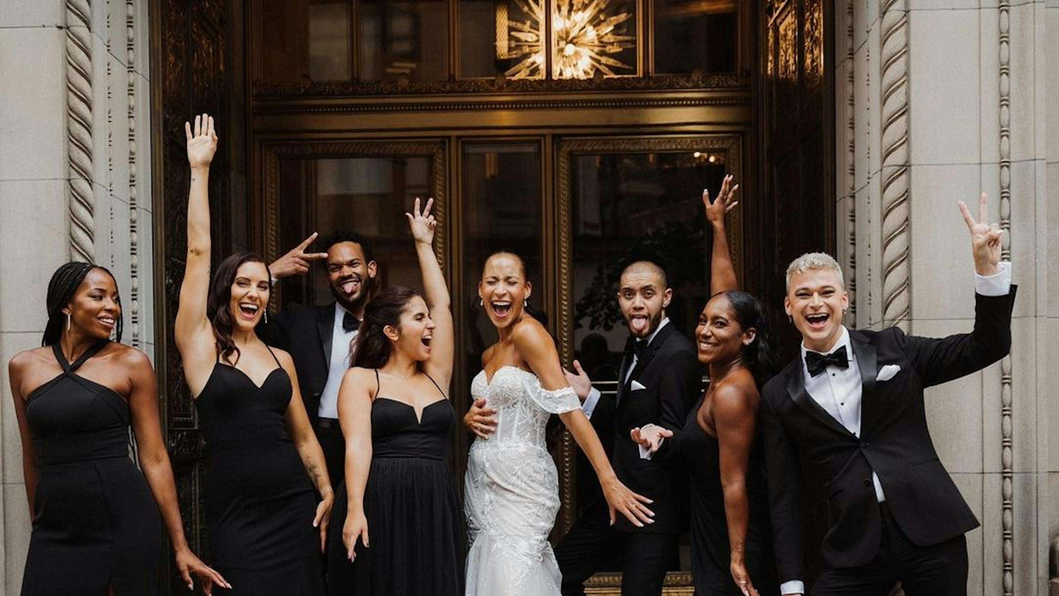All black bridal party outfit trend and black groomsmen tuxedos for a monochromatic black wedding party celebrating outside the wedding venue.