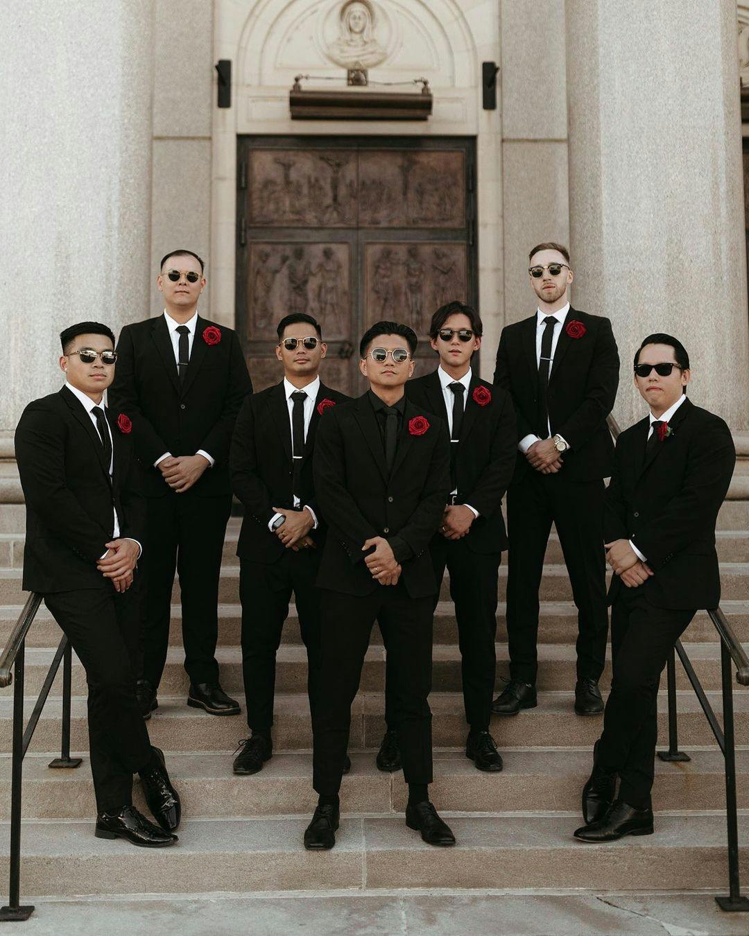 Black suit groomsman group with red rosette lapel pins and all-black suit ensemble for the groom.