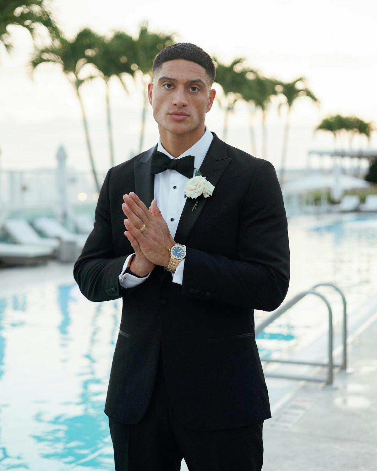 Man wearing summer black tie wedding style in a black tuxedo, metallic men's jewelry, and a white boutonnière by the pool with palm trees.