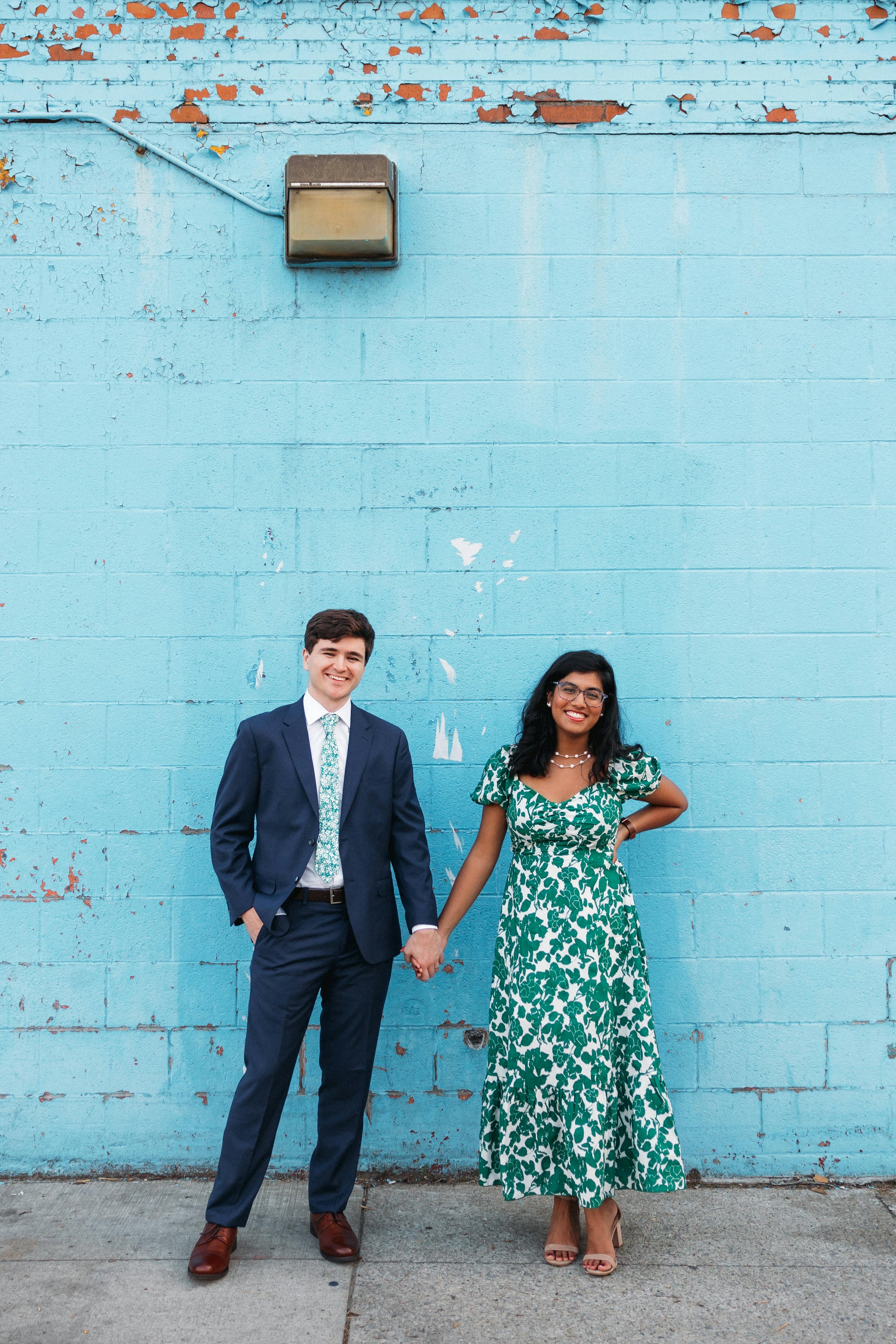 Groomsman in navy wedding suit with blue floral tie to match bridesmaid in green floral dress against bright blue photo backdrop.