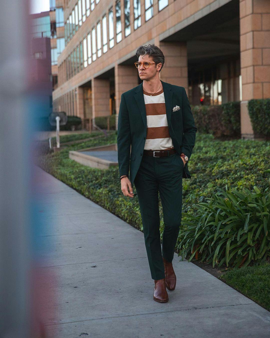 Unexpected way to style a suit professionally with a full dark green suit for men, contrasting rust orange striped t shirt, and brown suit accessories.