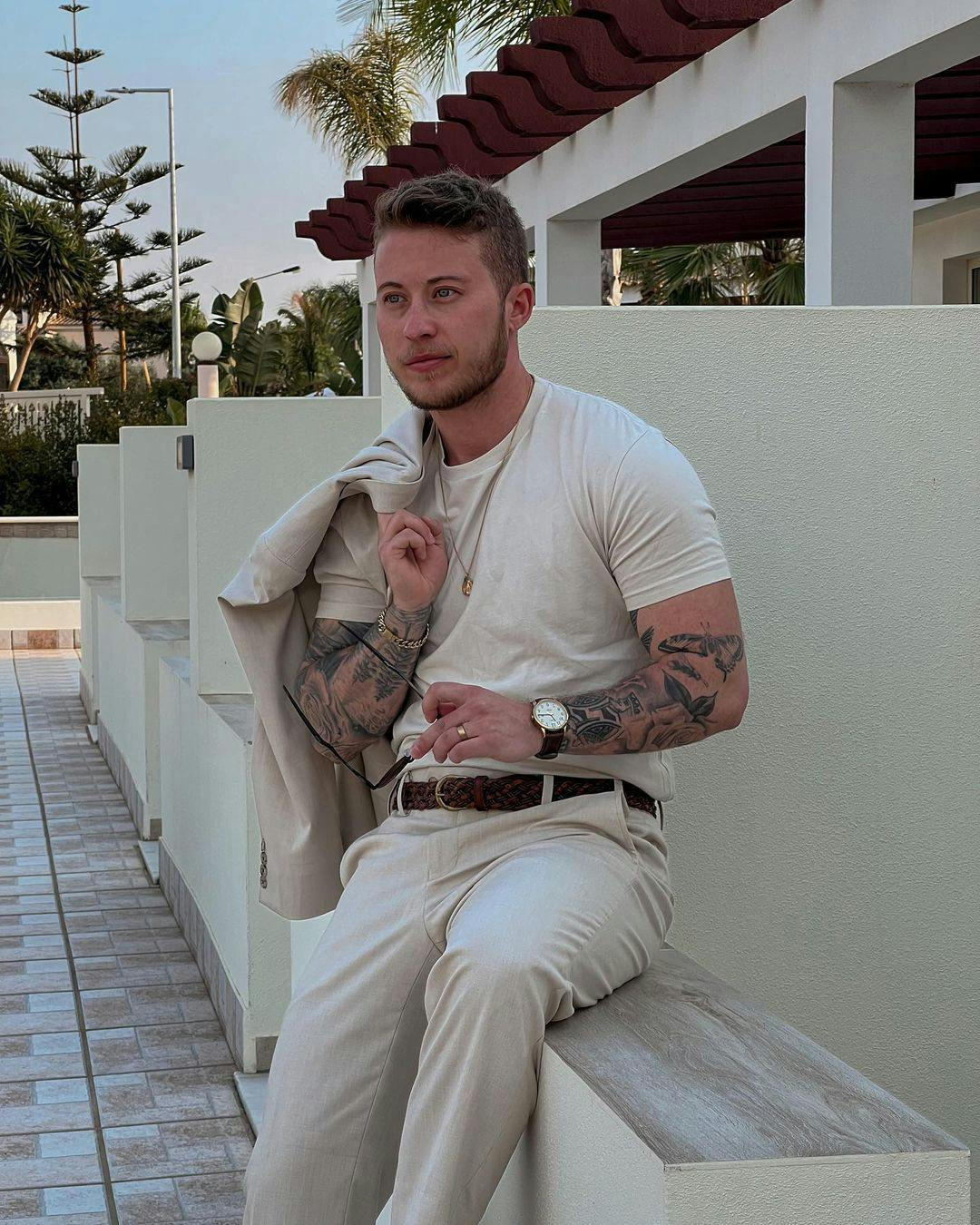 Man sitting outside wearing tan suit pants with a brown suit belt, beige t shirt, lots of men's accessories like a watch, chains, and glasses, and holding a light tan blazer.