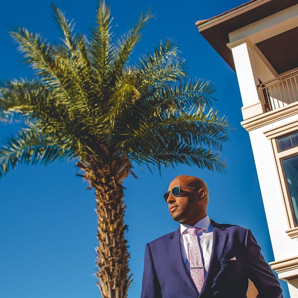 Easy beach wedding guest attire on a man in a navy blue suit, pink floral tie, matching pink pocket square, and sunglasses in front of a palm tree.