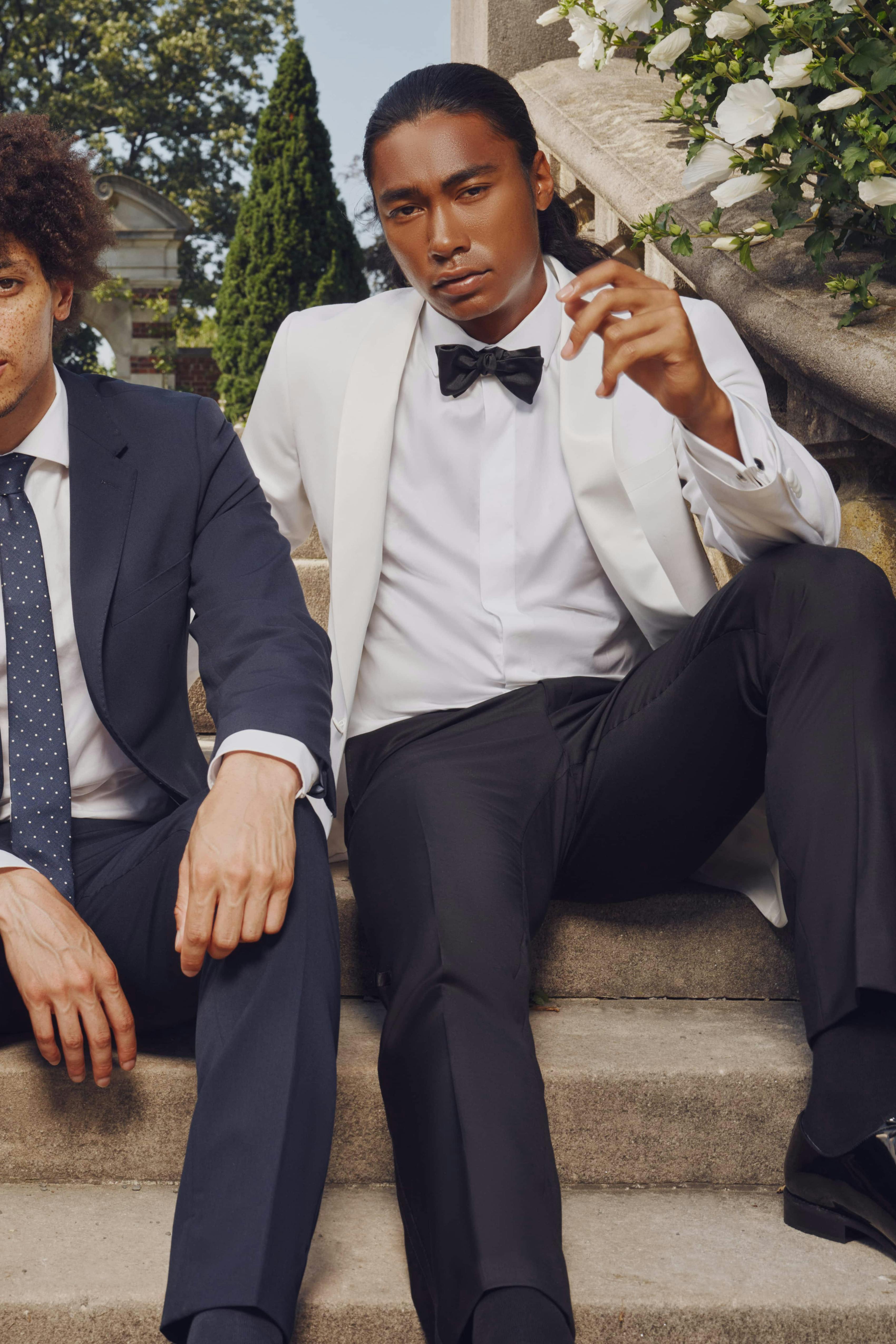 Wedding guest outfits of simple light blue suit and navy blue suit next to groom's white tuxedo at a garden party.