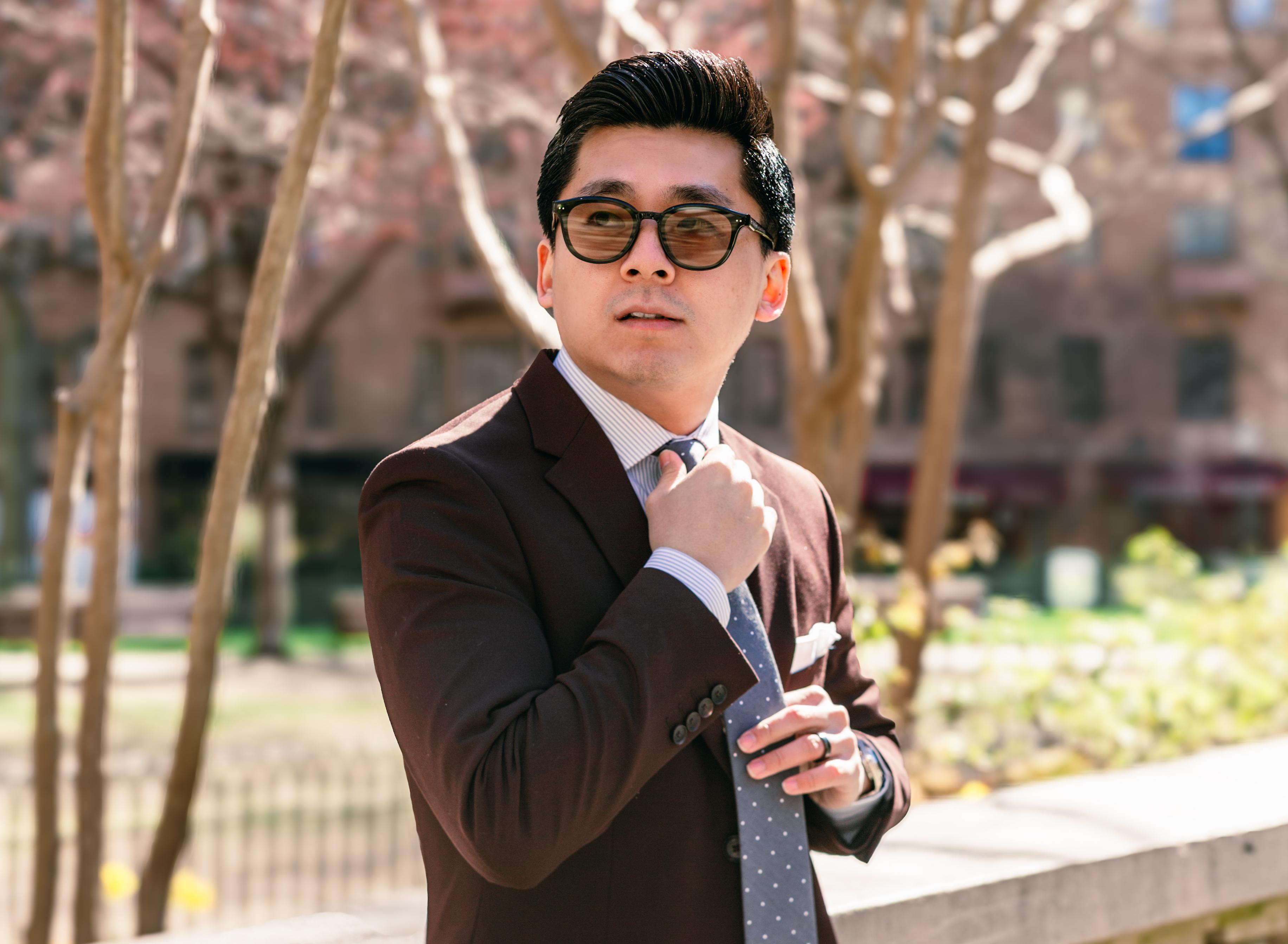 Man wearing a tailored dark brown suit, striped dress shirt, and navy polka-dot tie adjusting his tie while standing outdoors.