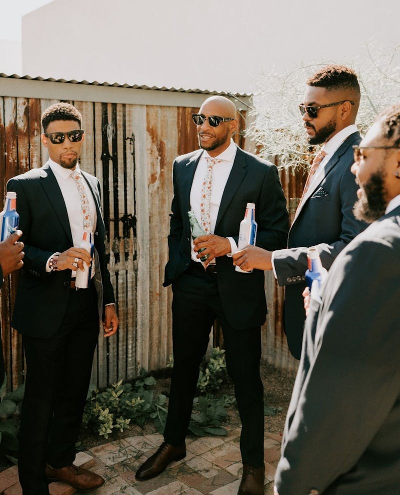 Groomsmen drinking beers in navy wedding suits with pink floral ties and sunglasses.