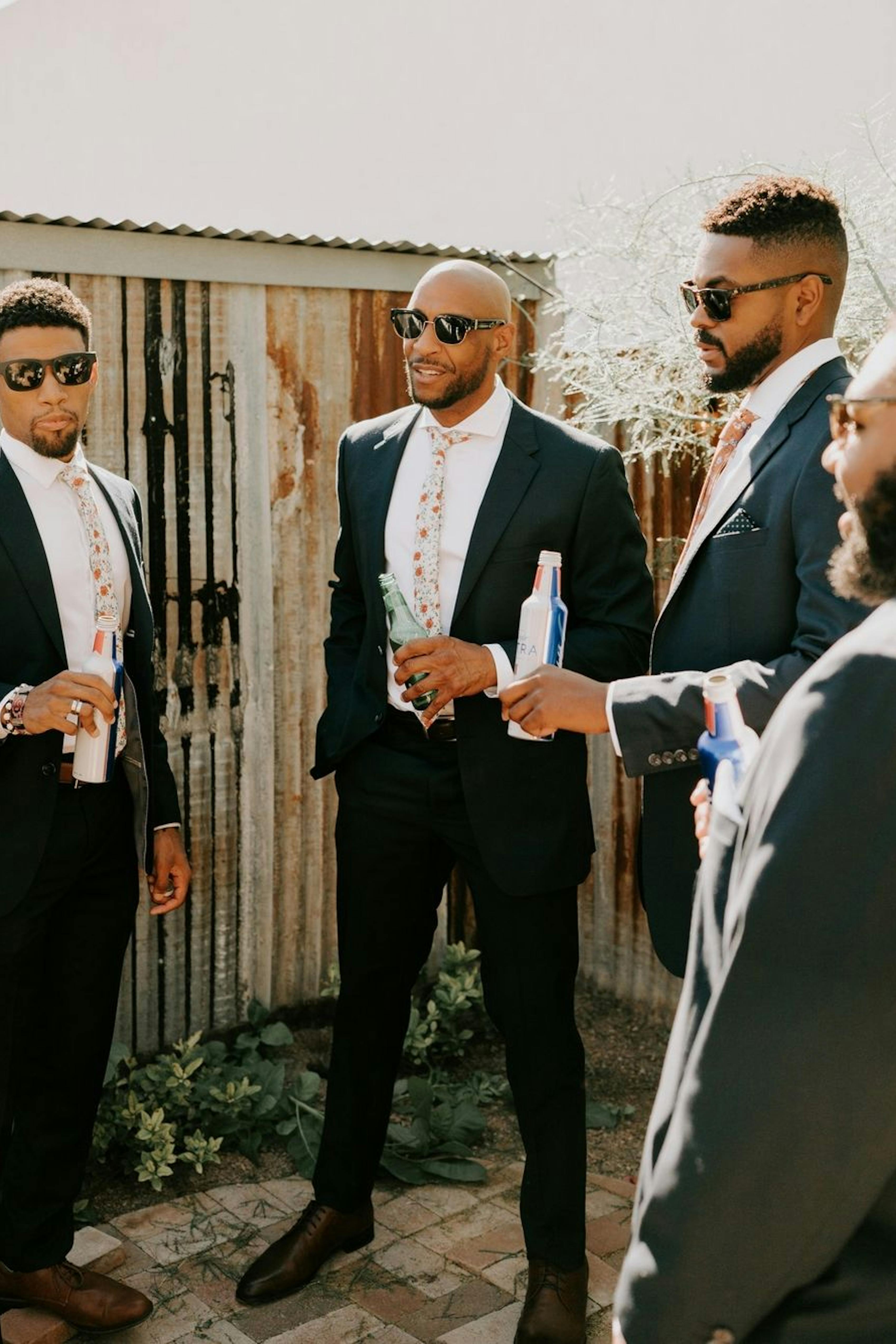 Groomsmen drinking beers in navy wedding suits with pink floral ties and sunglasses.