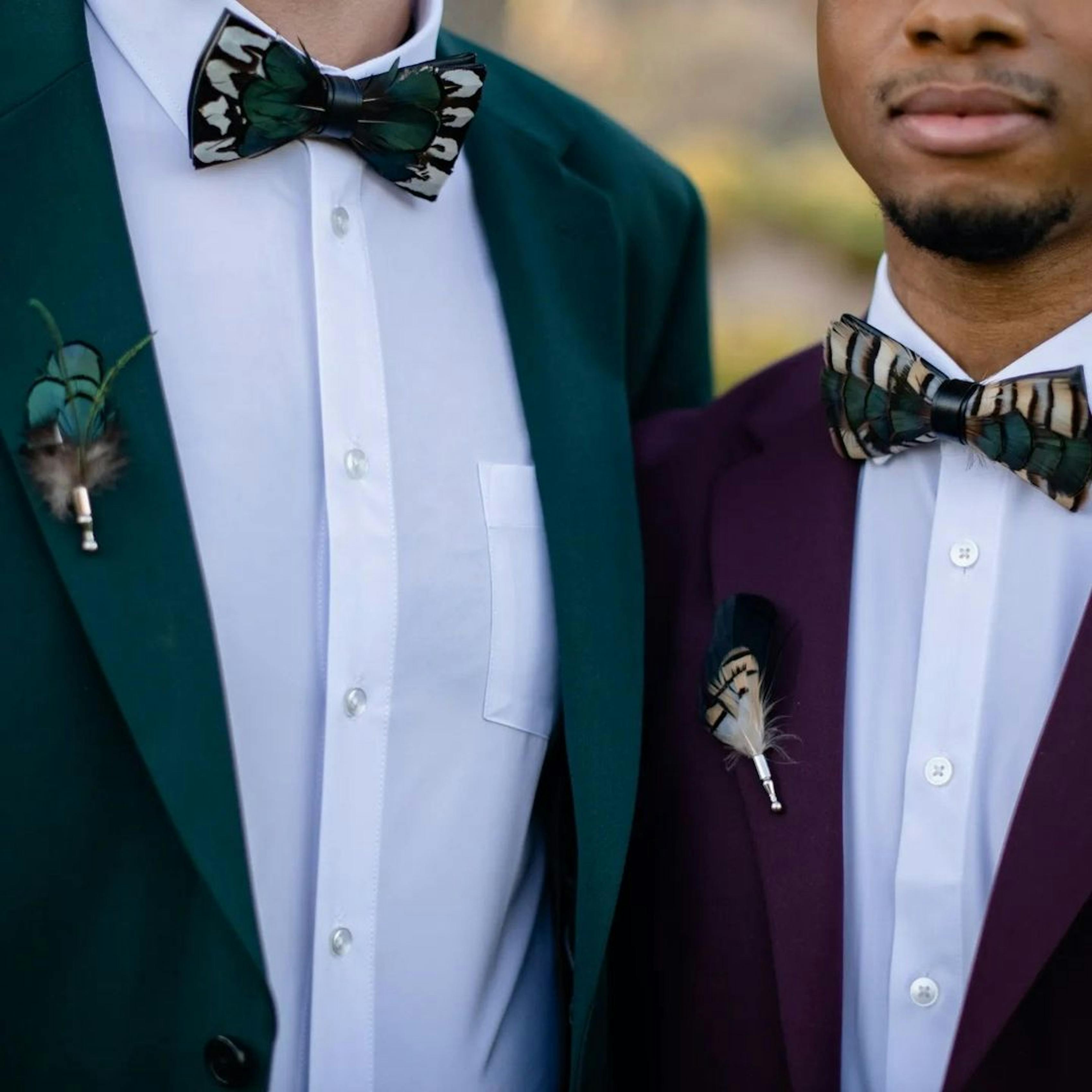 Gay couple wedding day suits with unique peacock feather bow ties and unique peacock feather boutonnières. Grooms wear men's dark green suit and men's burgundy suit.
