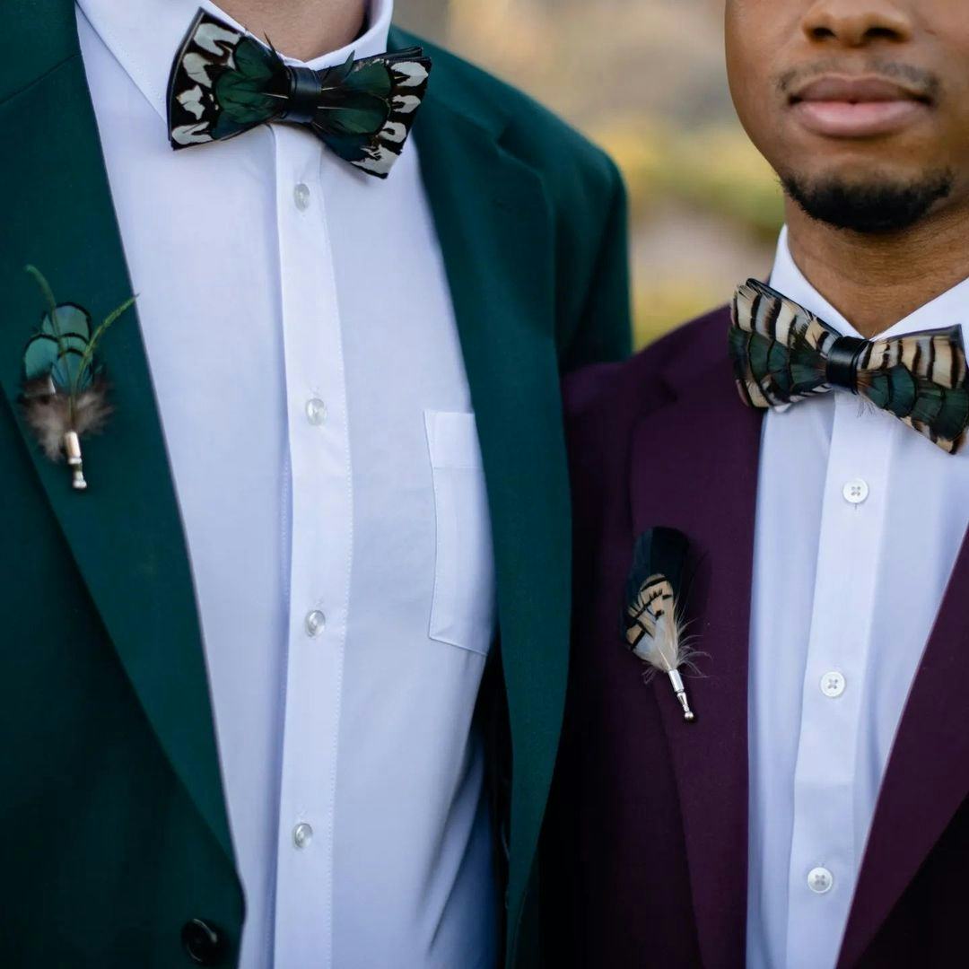 Gay couple wedding day suits with unique peacock feather bow ties and unique peacock feather boutonnières. Grooms wear men's dark green suit and men's burgundy suit. 
