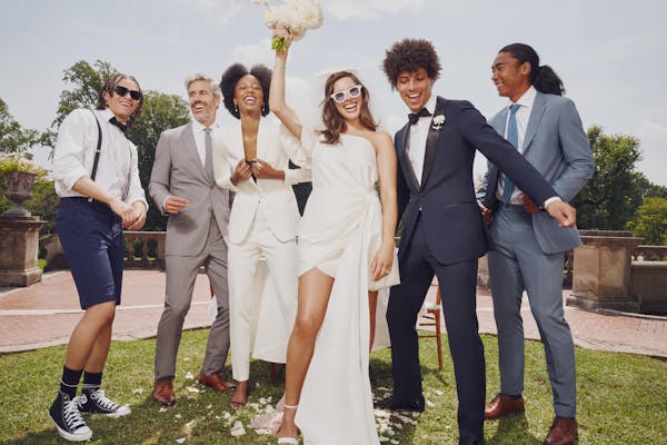 Wedding party of all ages with young teen groomsman wearing suit shorts, groom wearing navy tuxedo, bridesmaid wearing white suit, and wedding guests wearing formal suits at estate wedding