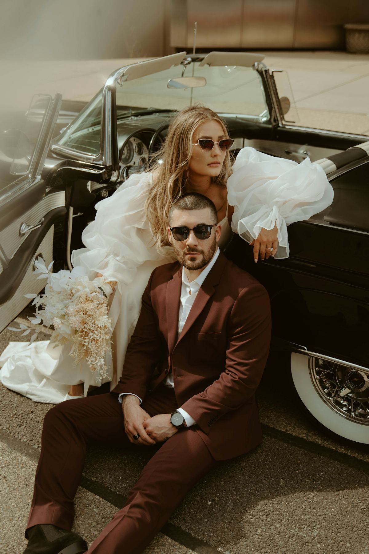 Alternative couple with an edgy wedding day sporting a maroon suit, dreamy, puffy sleeve dress posing with a vintage car.