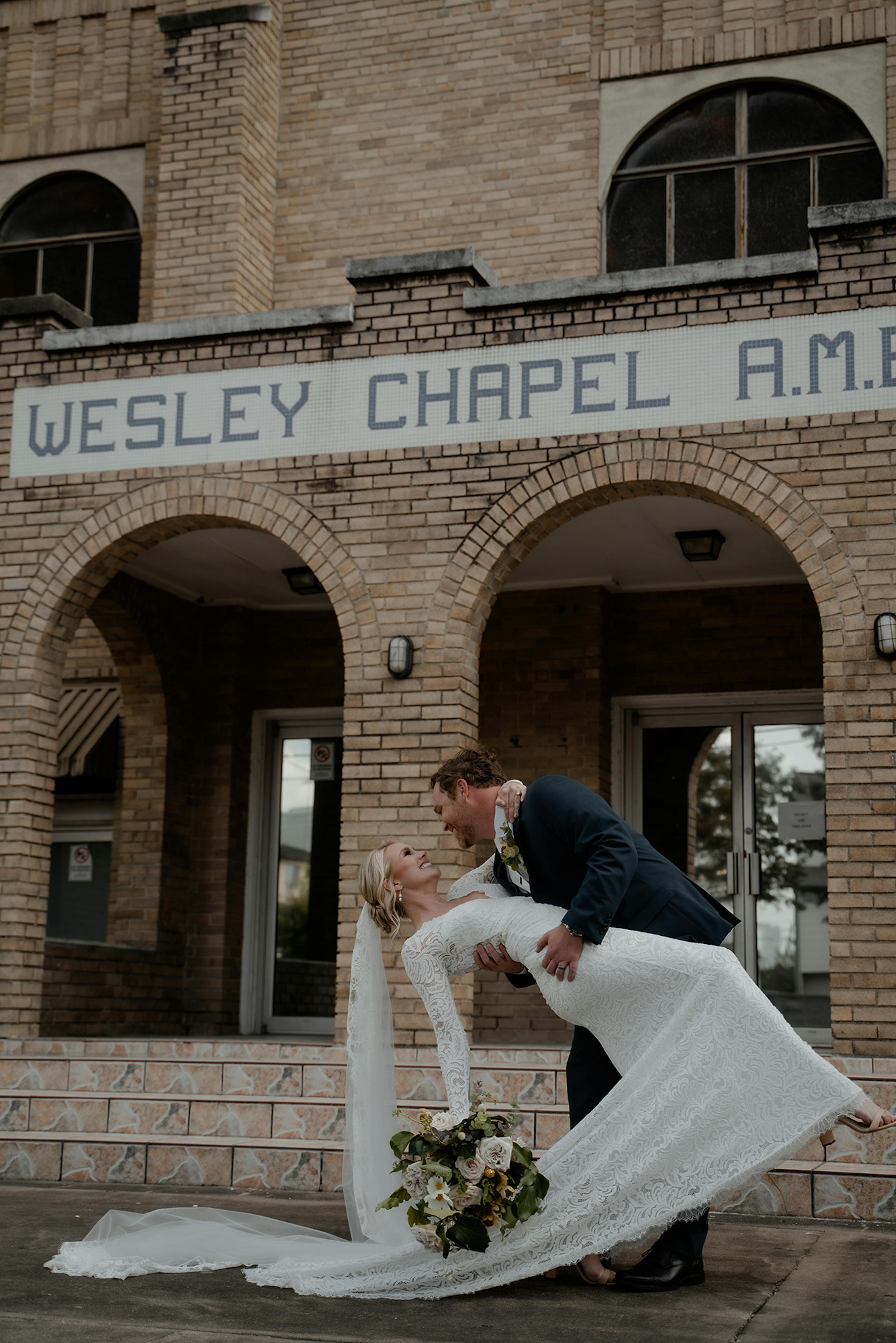 Just married bride and groom holding botanic bouquet wearing lace wedding gown and teal men's wedding suit.