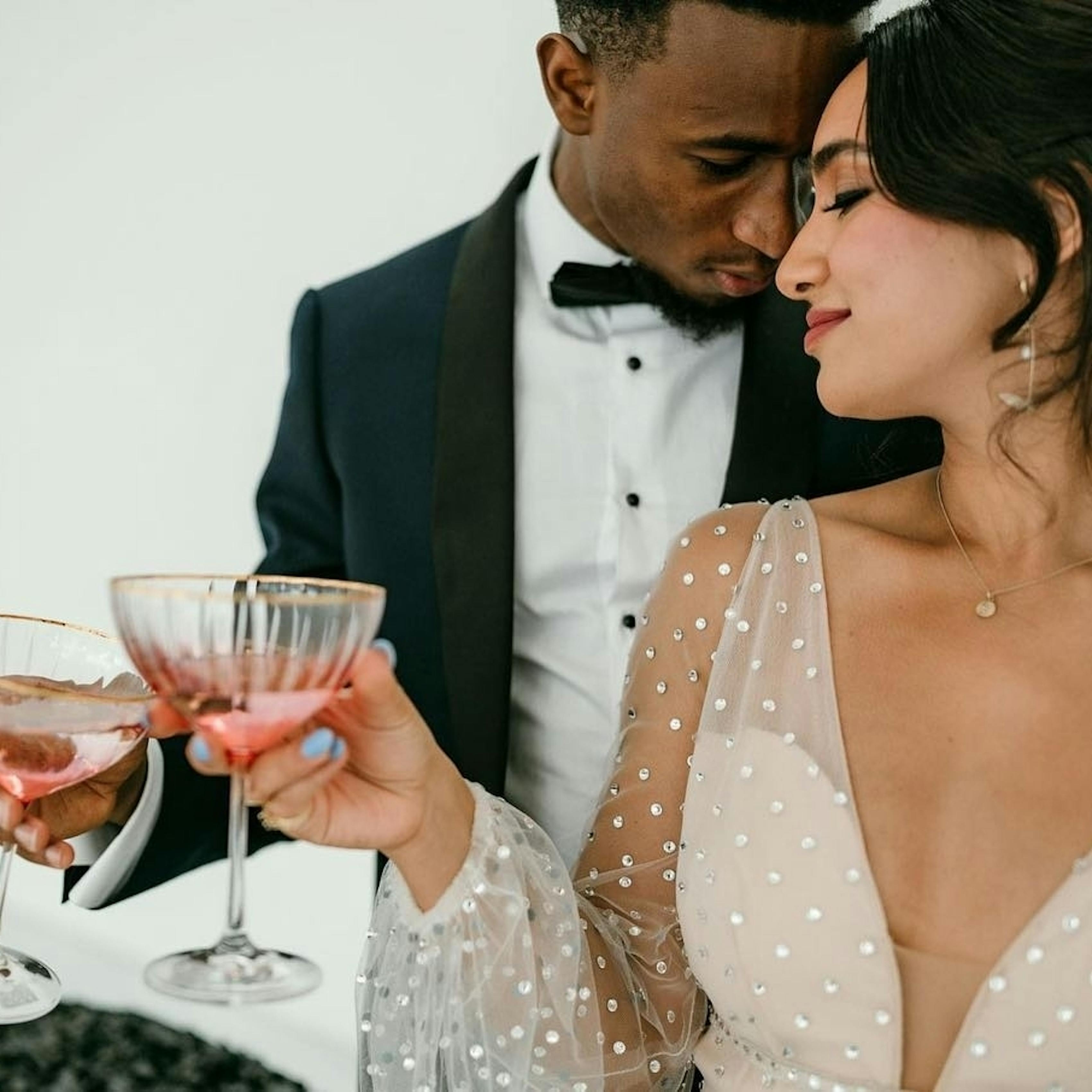 Bride wearing embellished sheer puff long sleeve wedding dress with bling and shine clinking glasses with groom in navy shawl lapel tuxedo for black tie wedding.