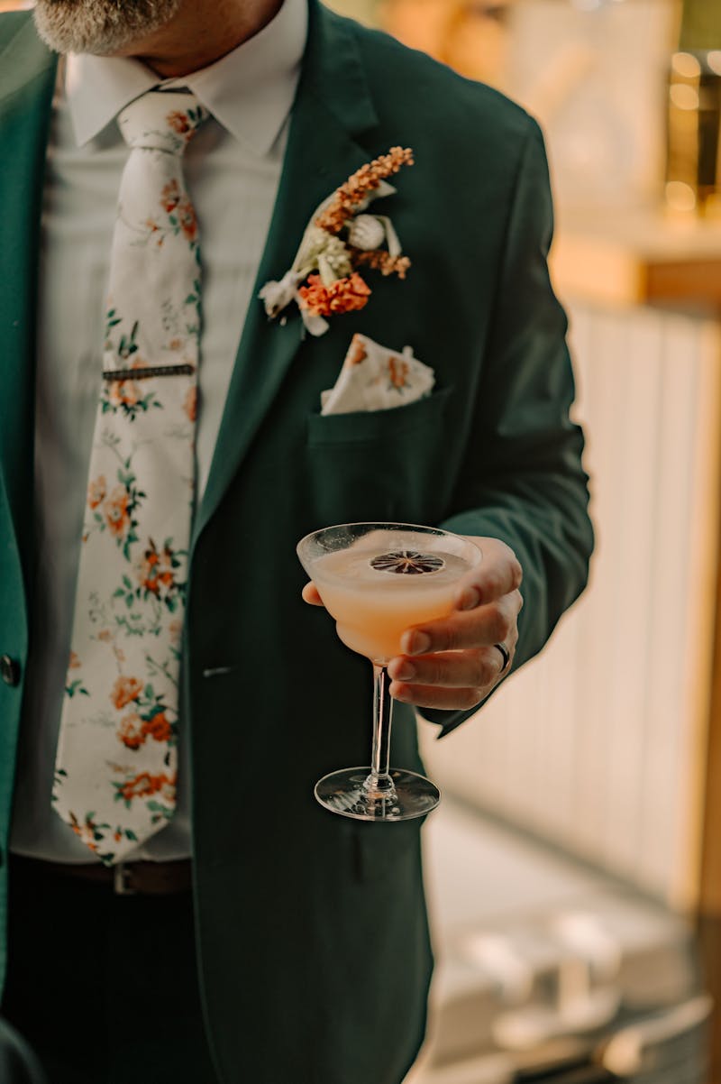 Father of the groom outfit ideas with man wearing dark green suit, floral tie, green and orange wedding color combination, and holding a signature cocktail.