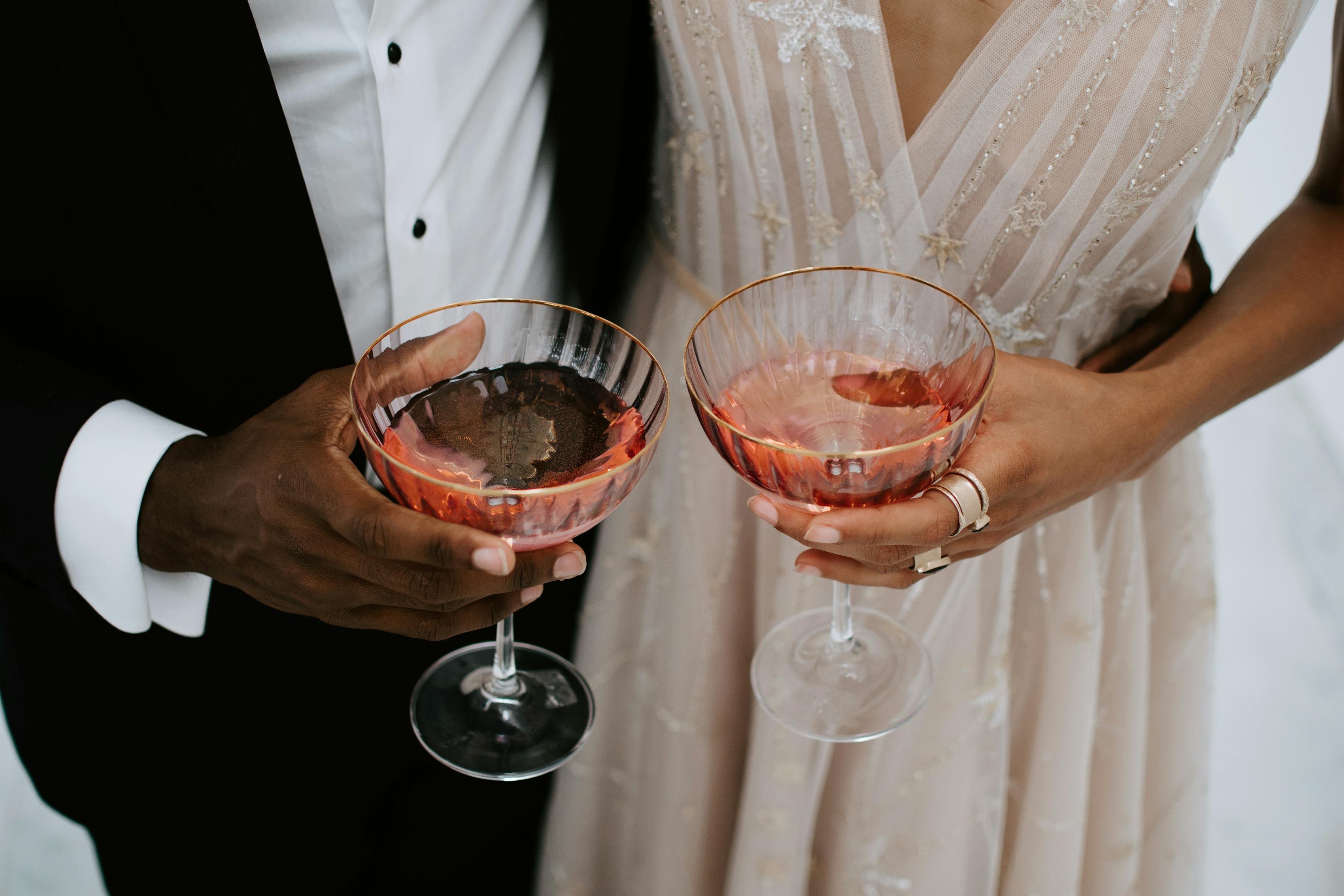 Bride and groom clinking glasses in champagne wedding dress with pink tones with classic black tuxedo