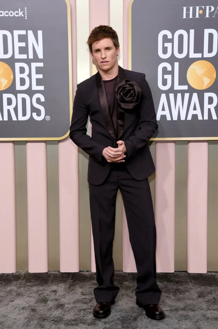 Eddie Redmayne wears an all black ensemble with a shawl lapel tuxedo, black shirt, and trending fabric flower on his lapel at the Golden Globes.