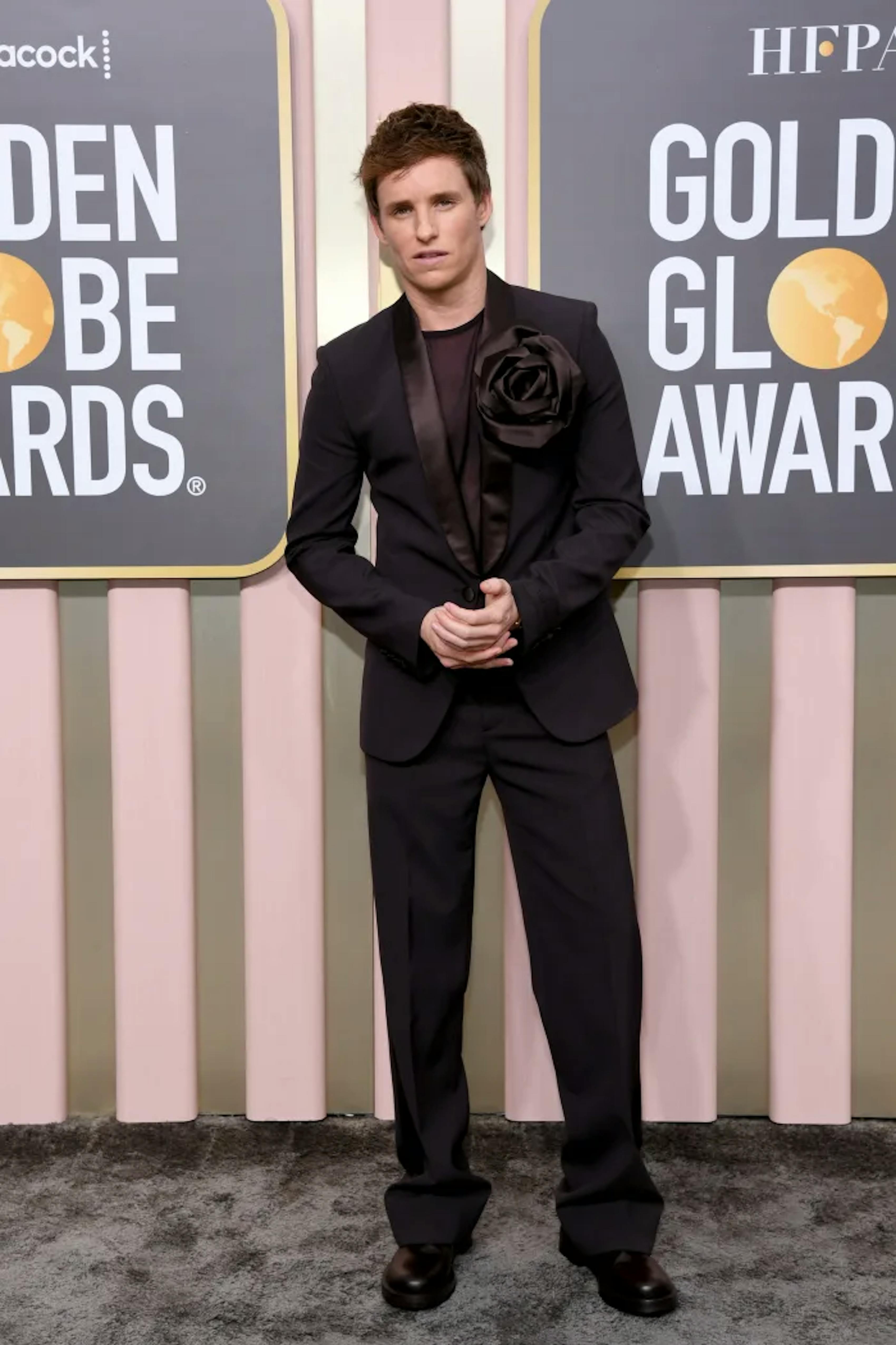 Eddie Redmayne wears an all black ensemble with a shawl lapel tuxedo, black shirt, and trending fabric flower on his lapel at the Golden Globes.