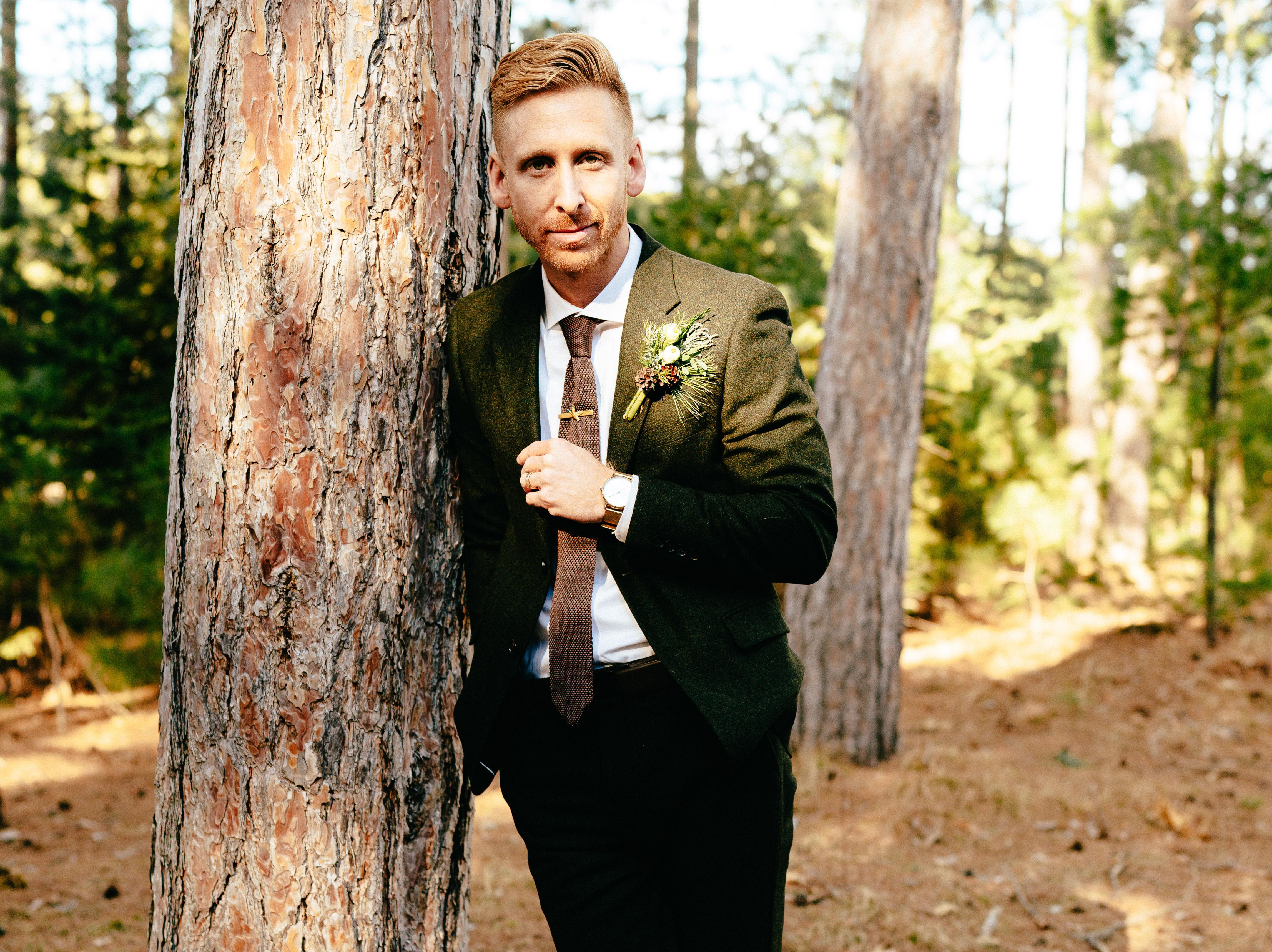 Man wearing an olive green suit jacket, brown tie, and floral boutonniere while leaning against a tree outdoors.