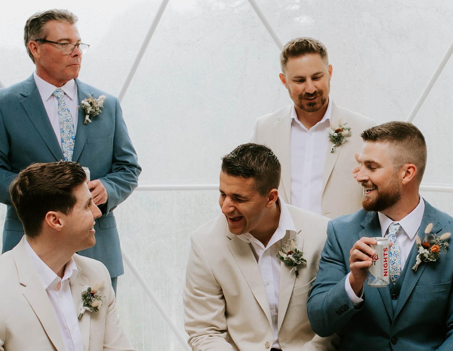 Five groomsmen laughing together in mismatched cream and dusty blue suits with floral boutonnieres at a wedding.