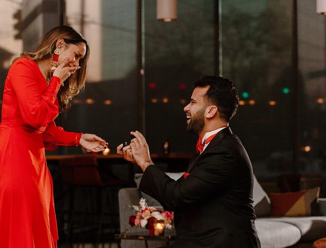 Man proposing on one knee to a surprised woman in a red dress during an engagement photo session.