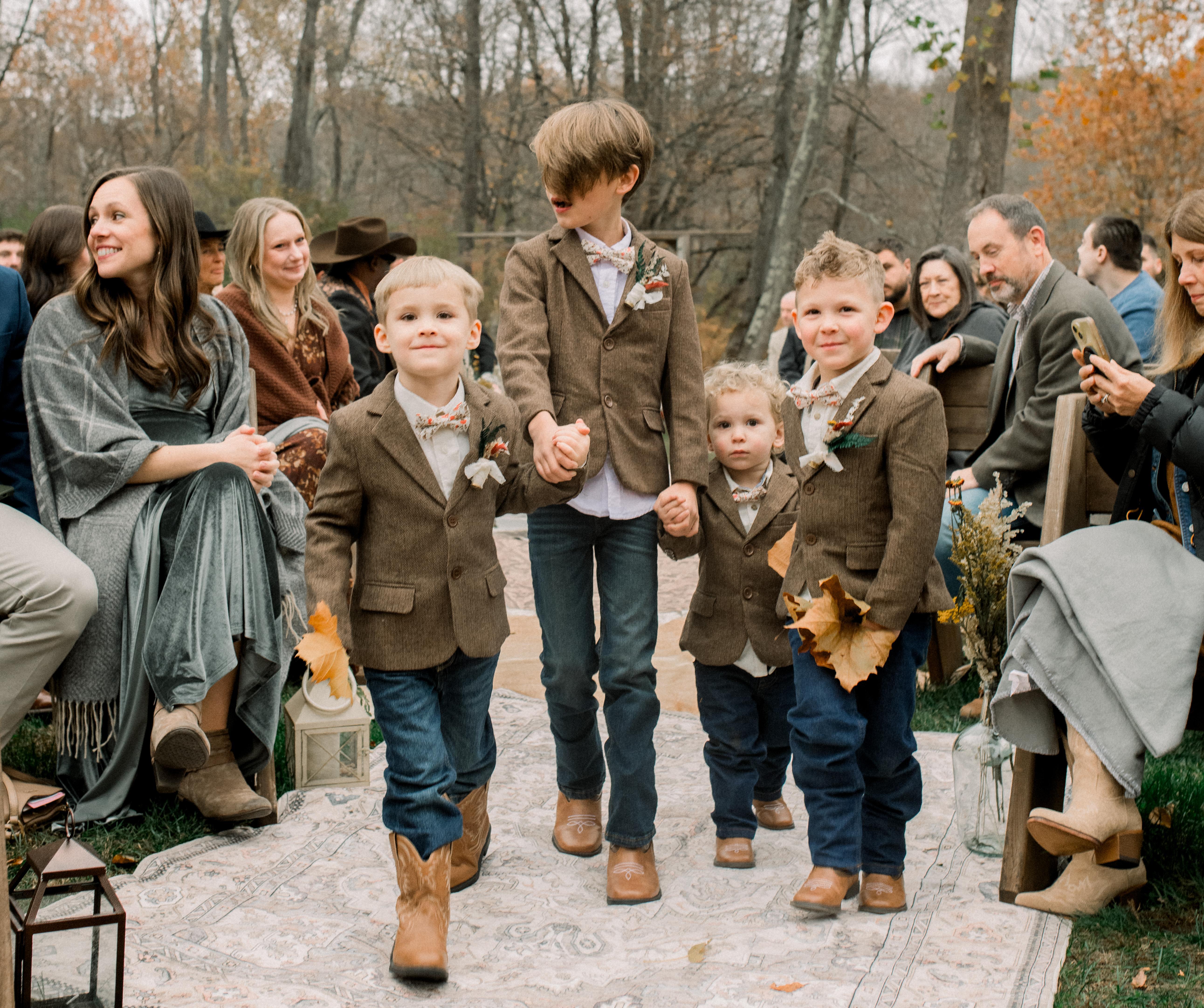 Children wearing brown jackets, bow ties, jeans, and boots walking down the aisle at an outdoor fall wedding.