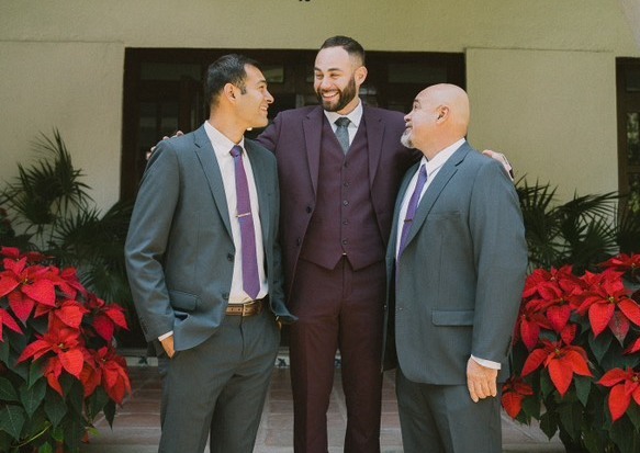 Three men wearing burgundy and gray suits pose together at a winter wedding in front of red poinsettias.