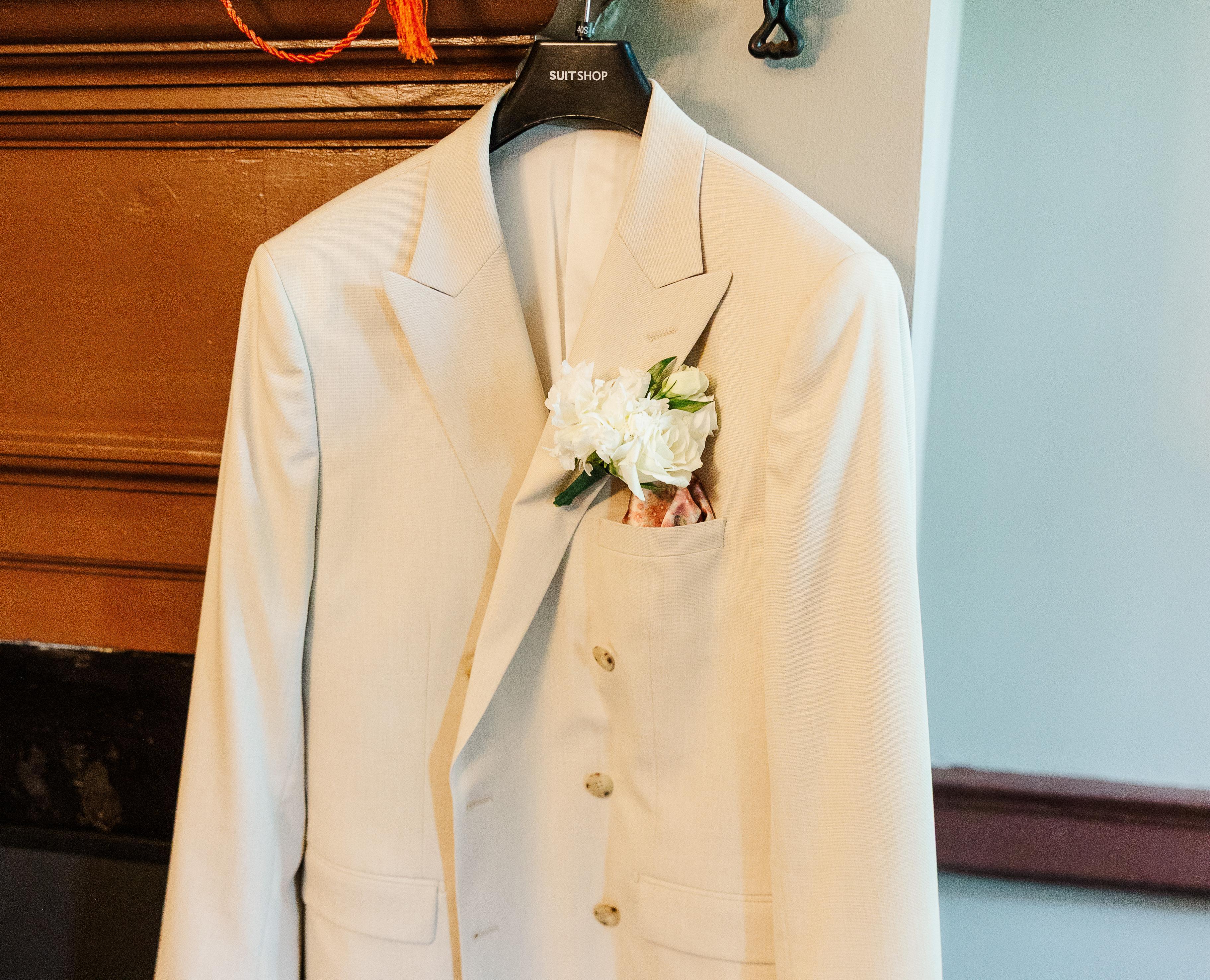 White wedding suit jacket with floral boutonniere displayed on a hanger.