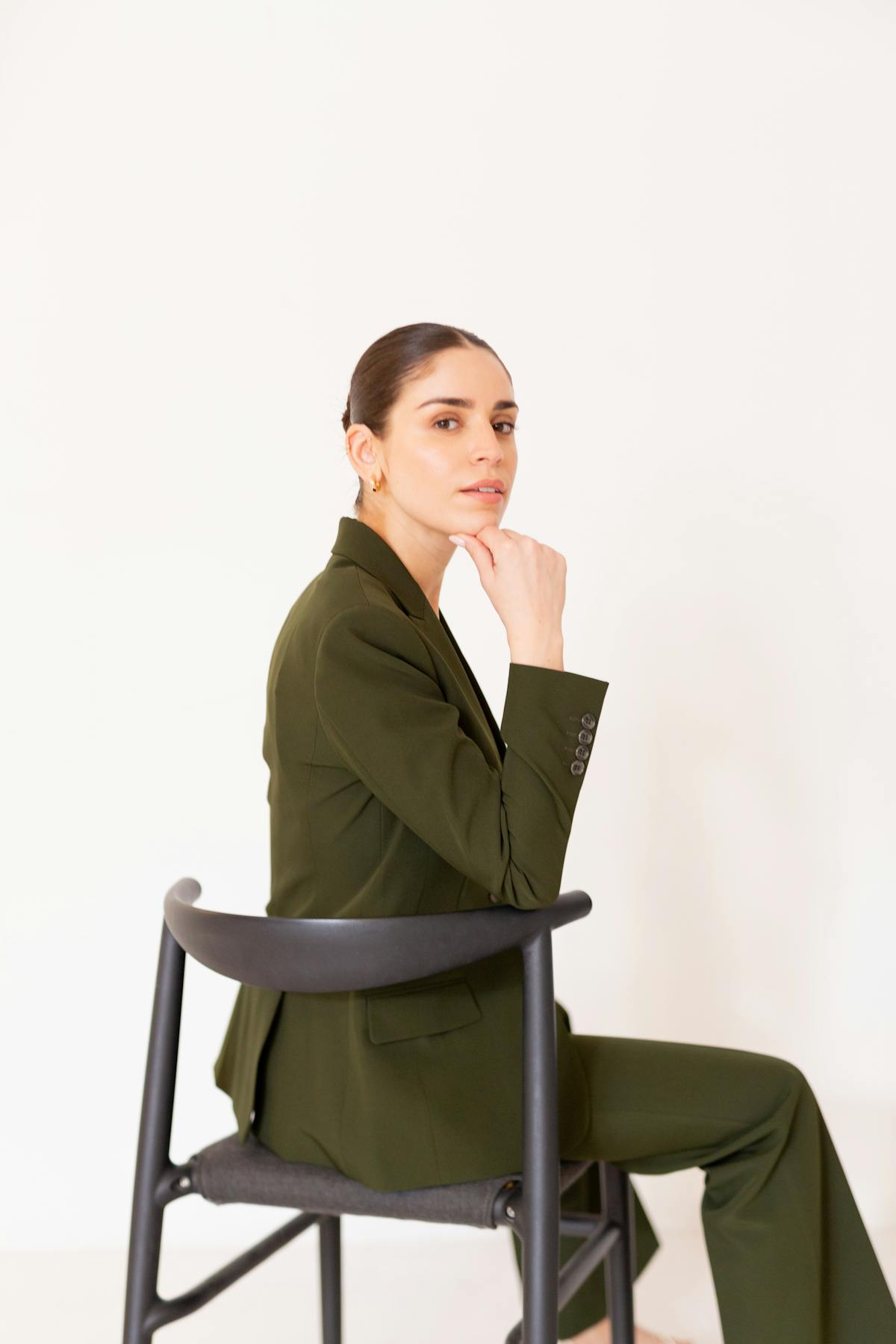 Woman wearing olive green pantsuit outfit sitting in chair looking thoughtful