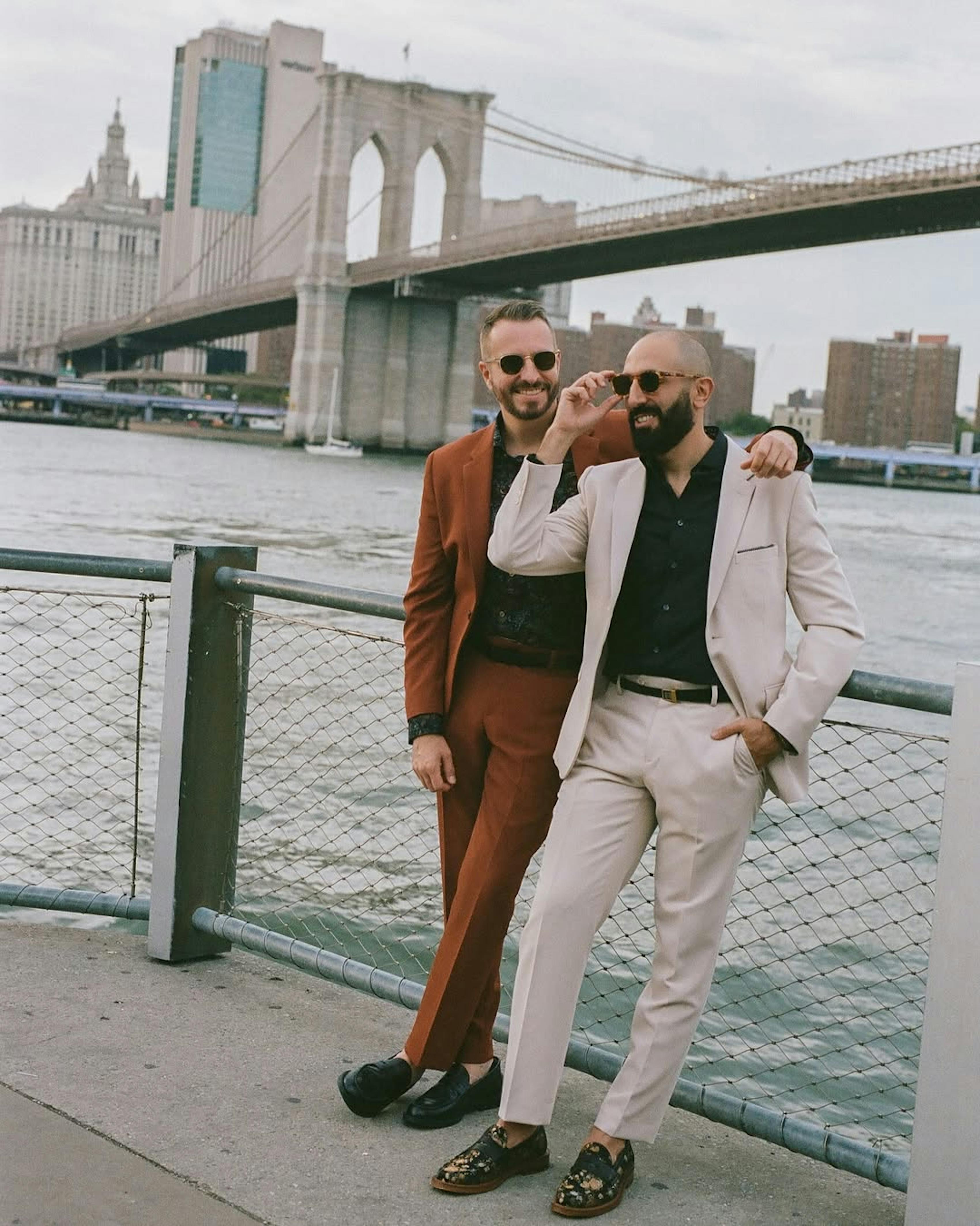 Smiling wearing men's light tan suit and men's brown clay suit with black dress shirts in front of Brooklyn Bridge