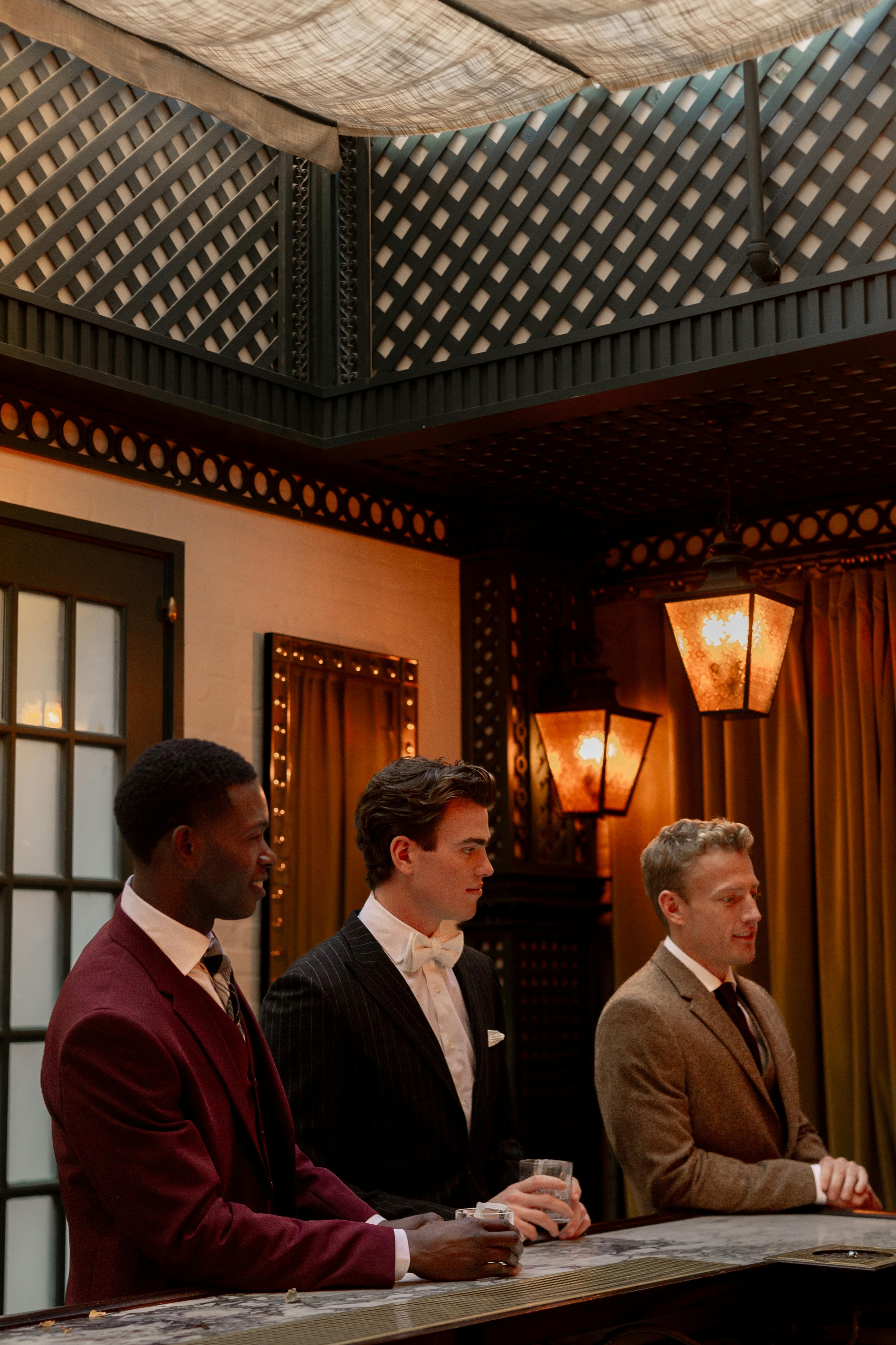 3 men at wedding bar wearing merlot suit, black pinstripe suit, and tweed suit