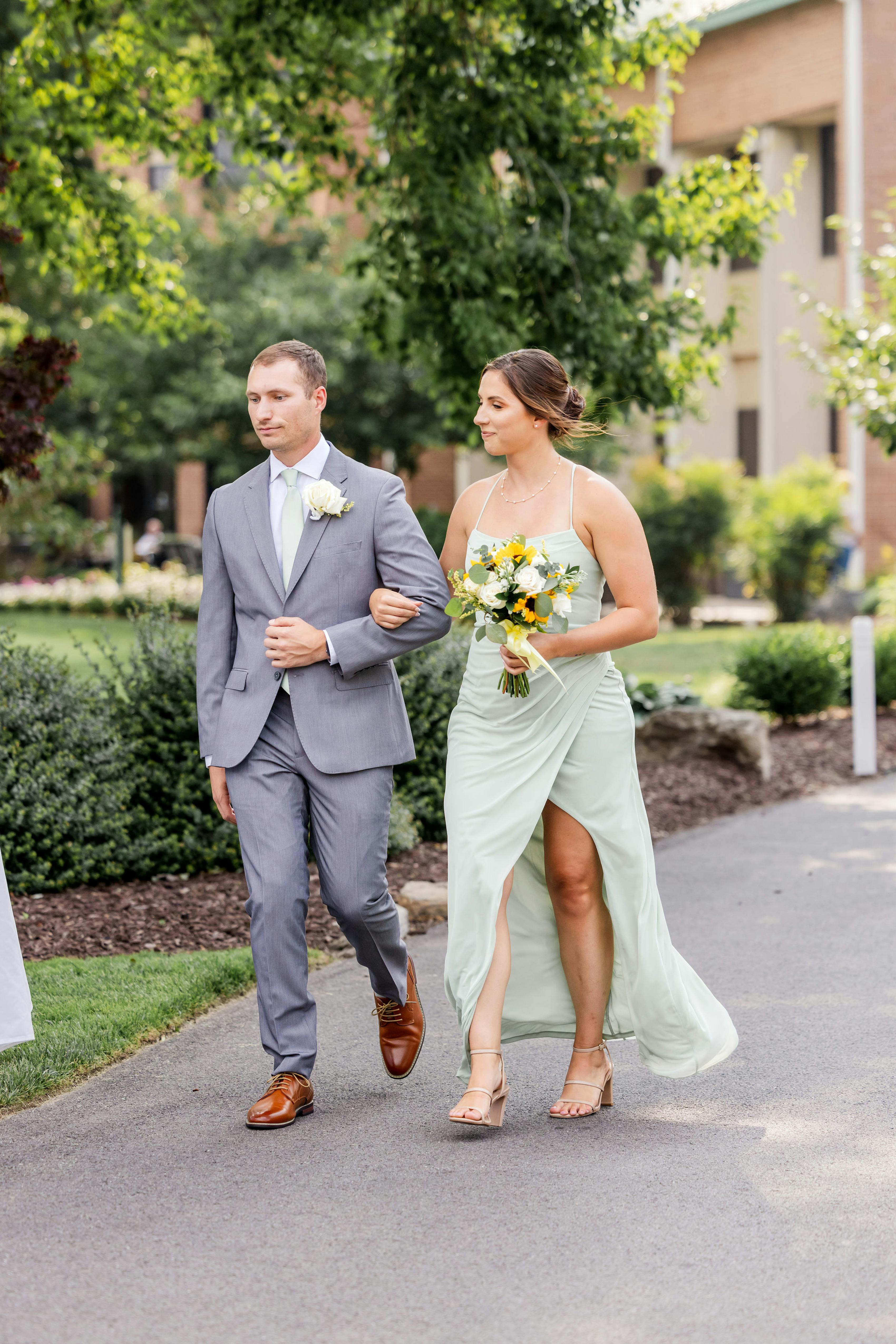 Sage green bridesmaid dress and light grey suit match walking down aisle