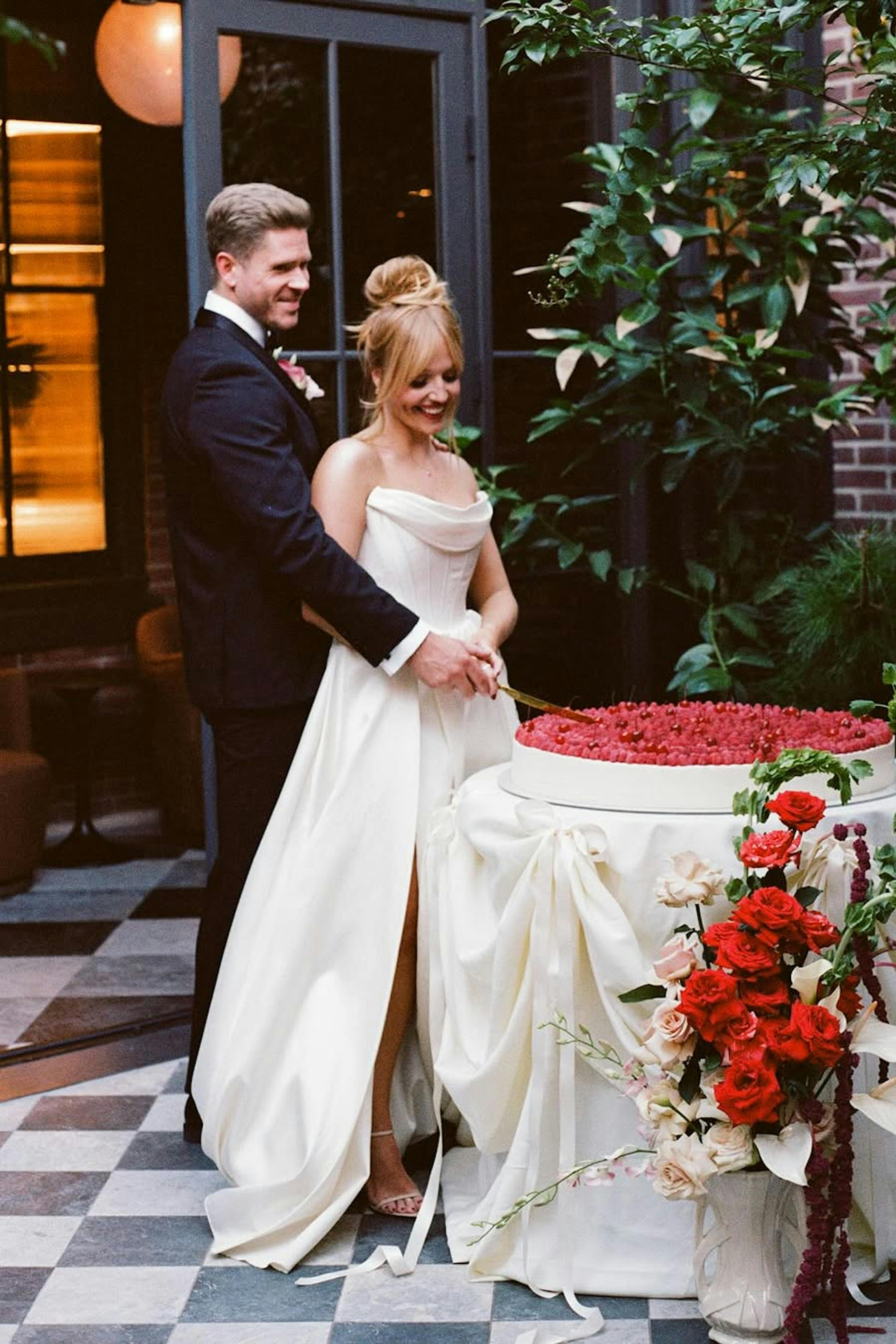 Cherry covered wedding cake cutting with red florals and black tie tuxedo and gown