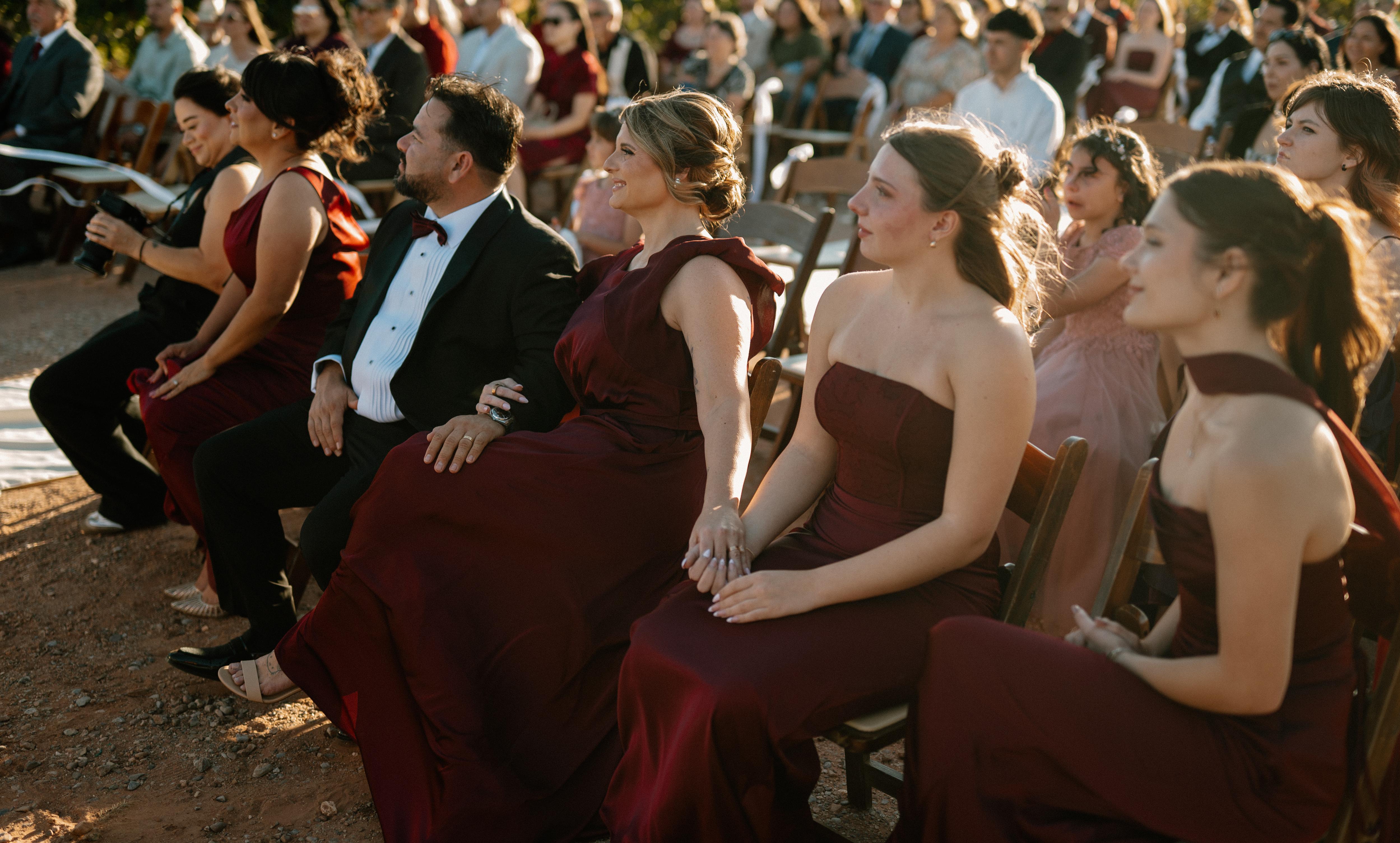 Winter wedding guests seated outdoors, wearing burgundy dresses and a black tuxedo with a burgundy bow tie during the ceremony.