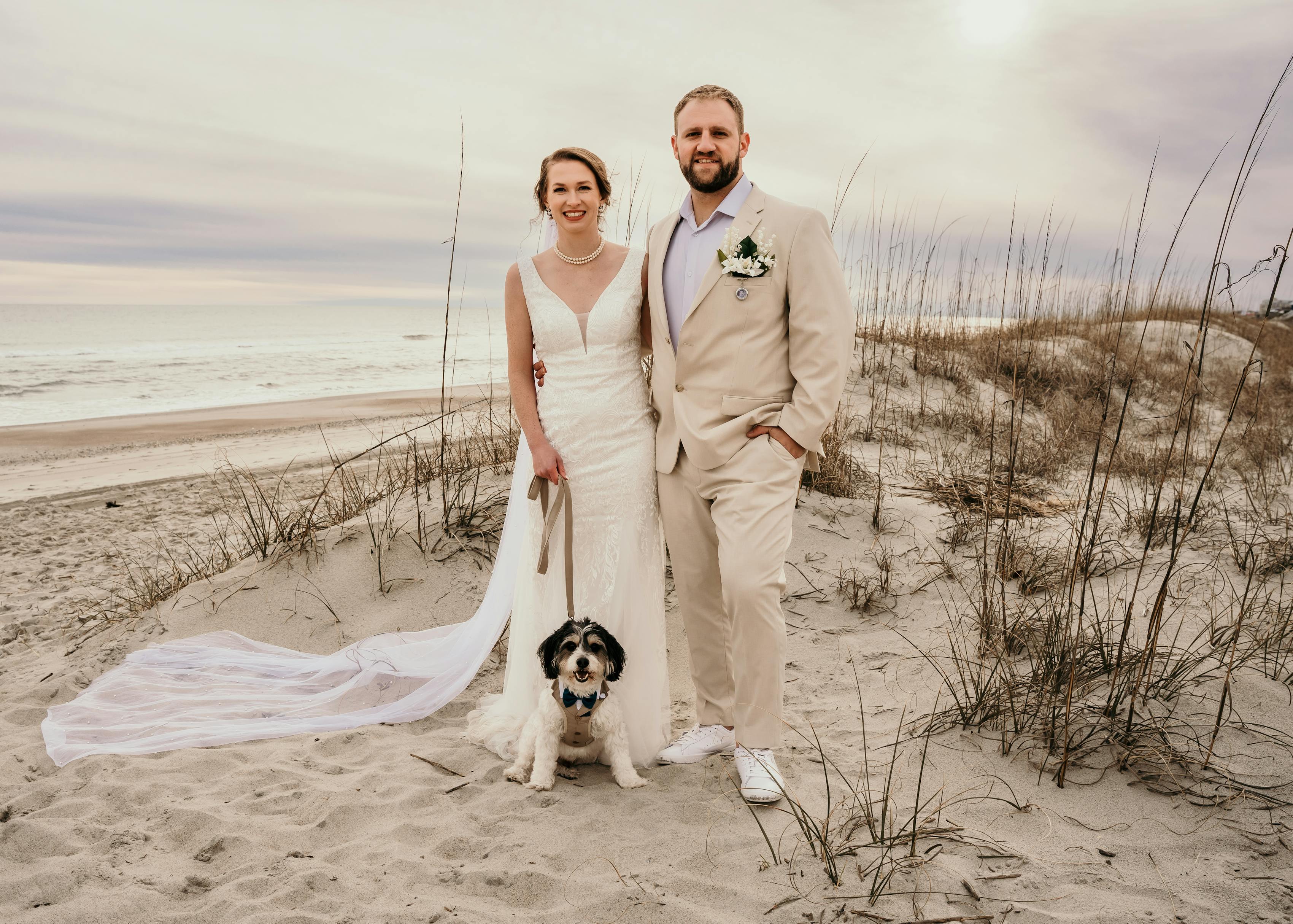 bride and groom pose beachside with their dog in their neutral wedding attire