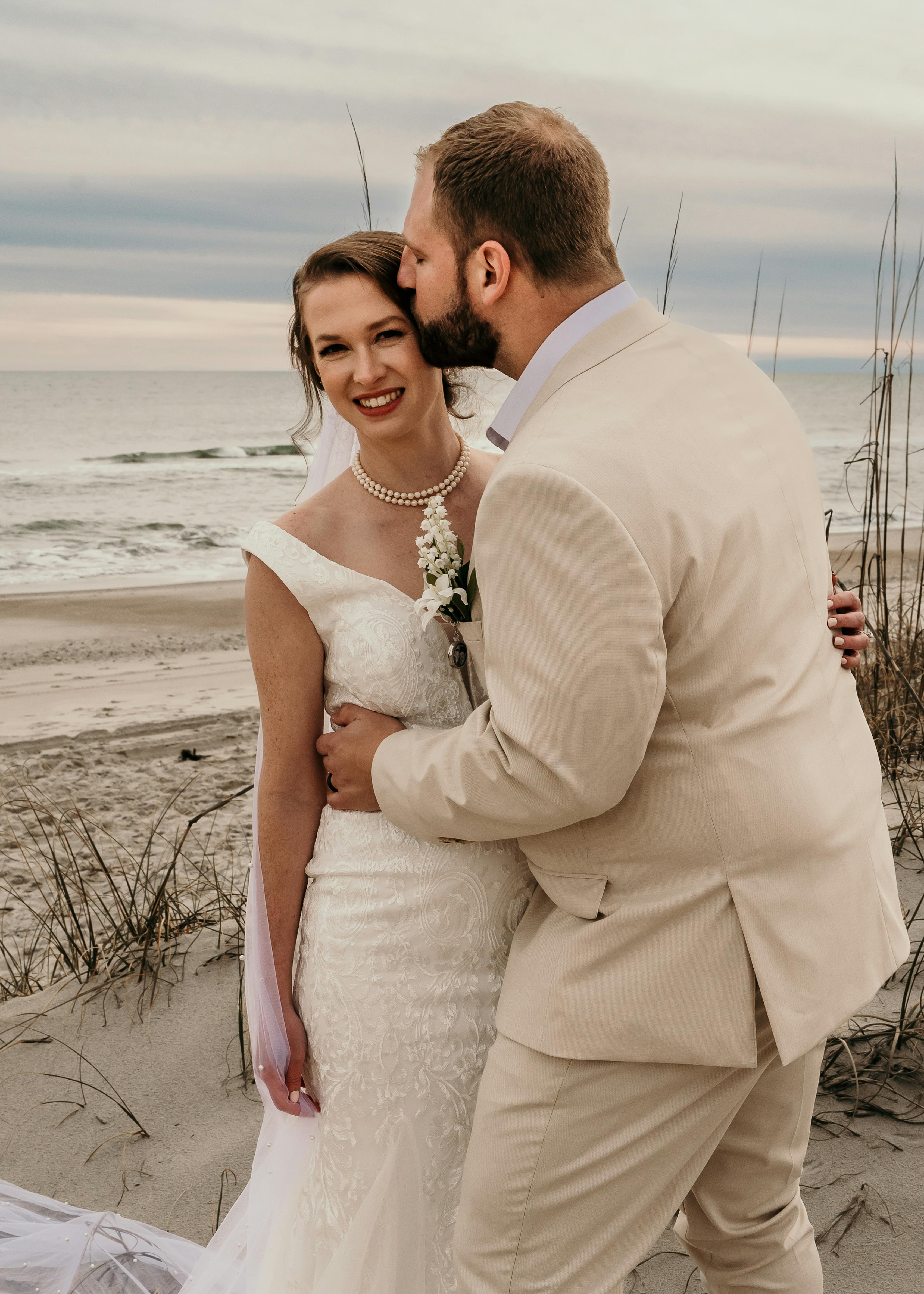 groom in tan wedding suit kissing bride on the beach