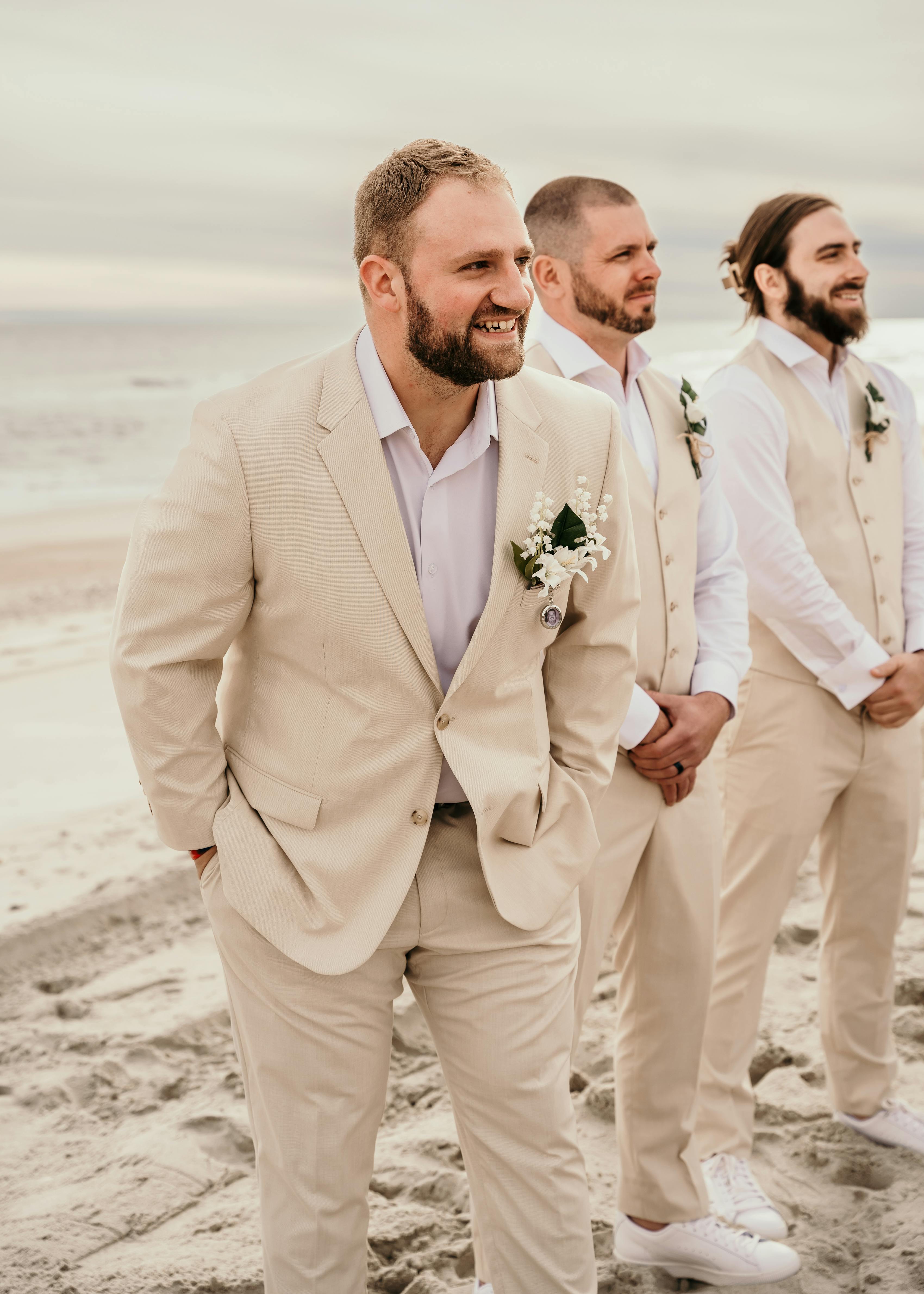 groom in tan suit stands next to groomsmen in tan vests and sneakers on the beach