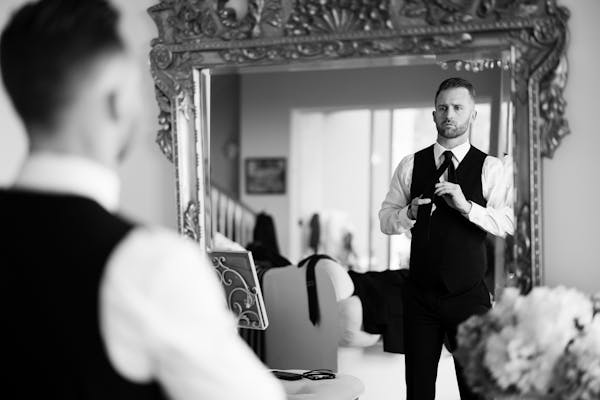 Man getting dressed to be a wedding guest tying neck tie in the mirror to match his black suit vest and suit pants.